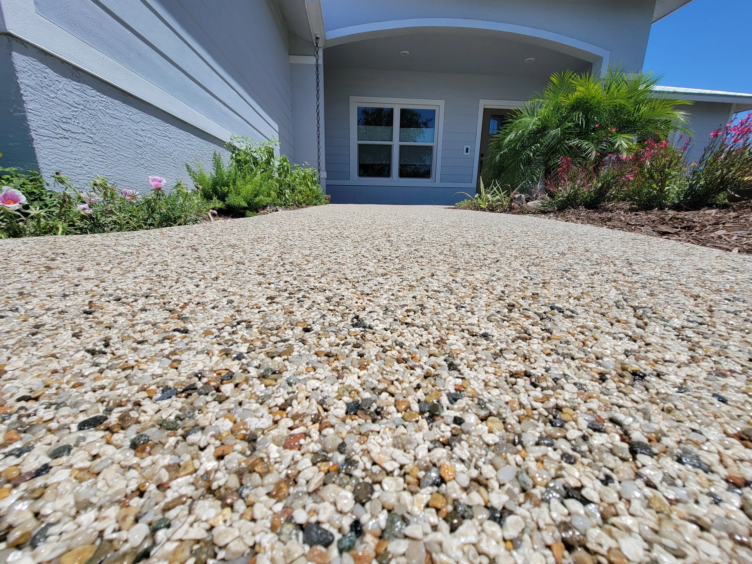close-up view of textured concrete driveway leading to the front door of a house with landscaping on both sides, including green bushes and pink flowers, under a clear blue sky