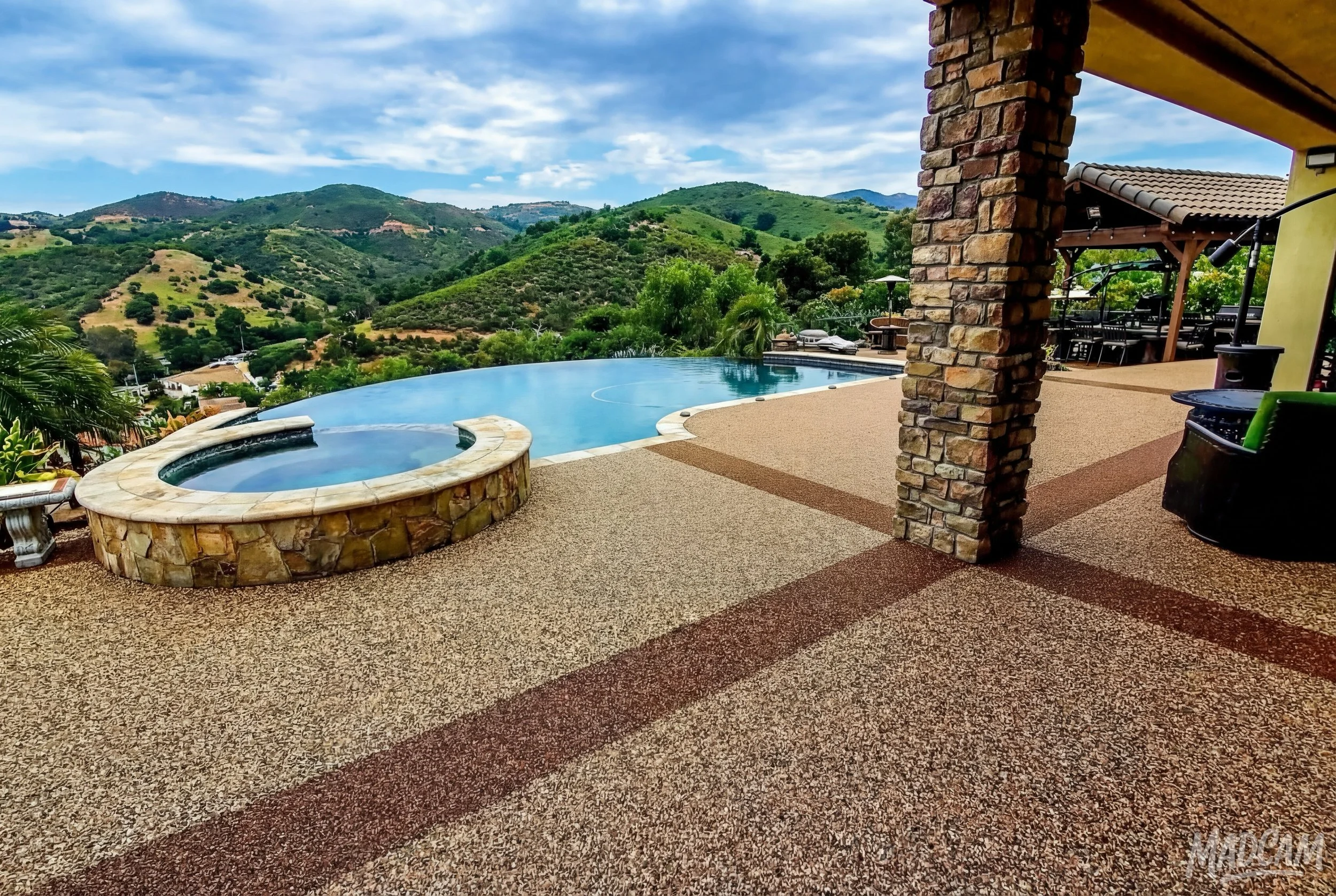 Outdoor terrace with a hot tub and infinity pool overlooking mountainous landscape with green hills and a partly cloudy sky.