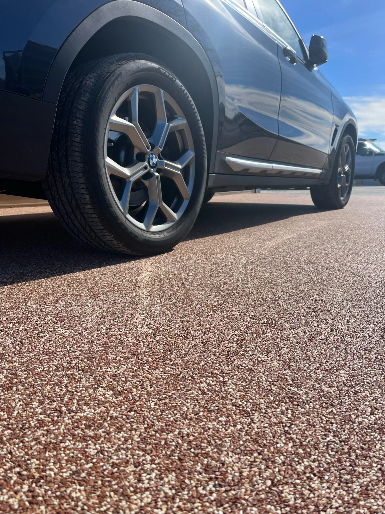 Close-up of a black BMW SUV parked on a textured parking lot surface with a blue sky overhead.