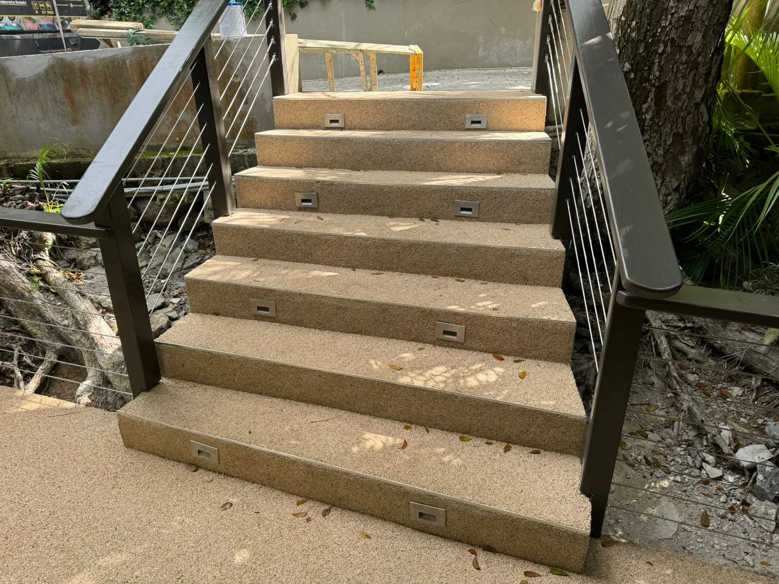 Outdoor staircase with six steps, black metal handrails on both sides, small rectangular lights embedded in each step, surrounded by trees and greenery.