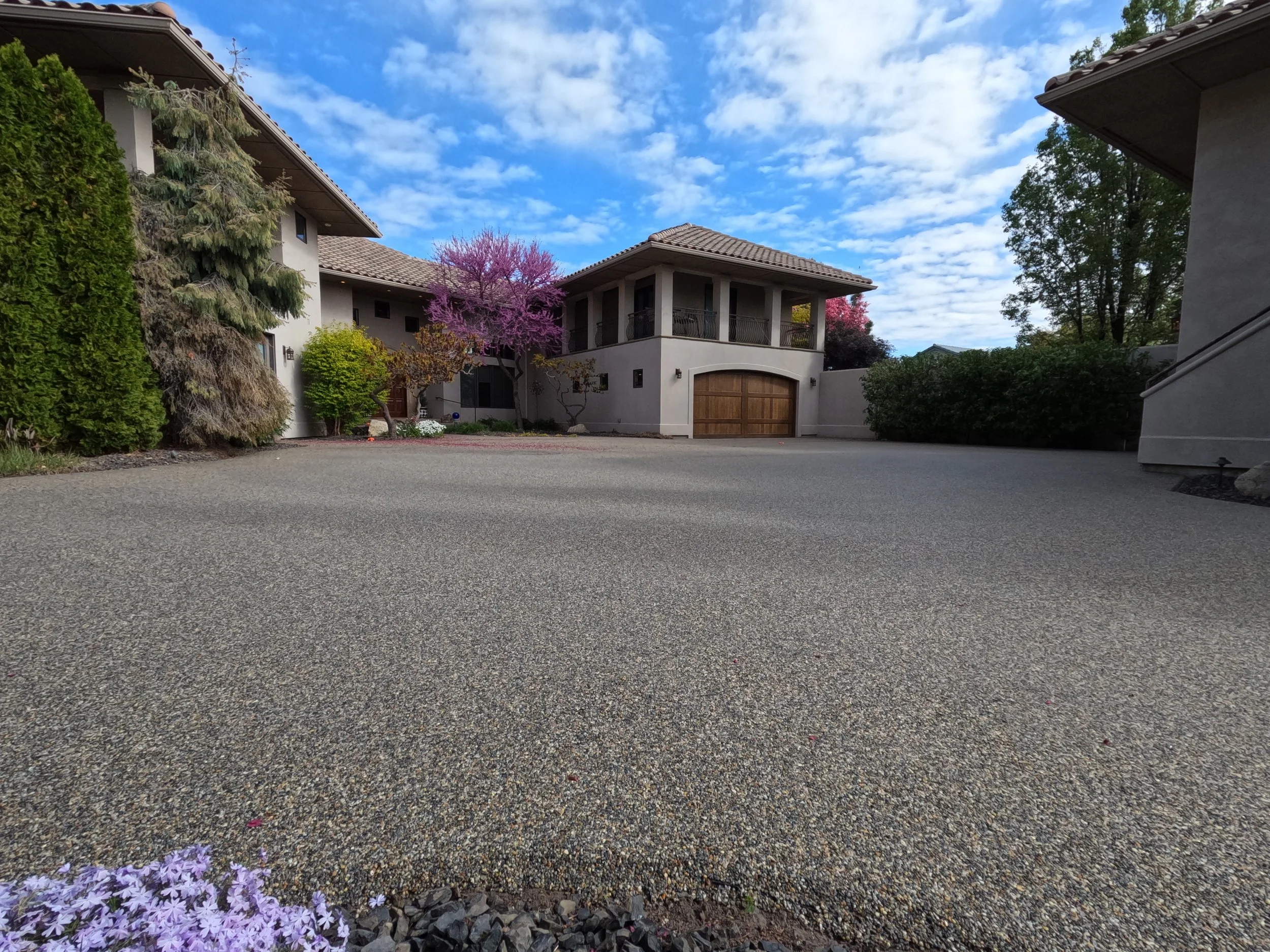 View of a large, paved driveway in front of a modern residential house with multiple levels, surrounded by greenery and trees under a partly cloudy sky.