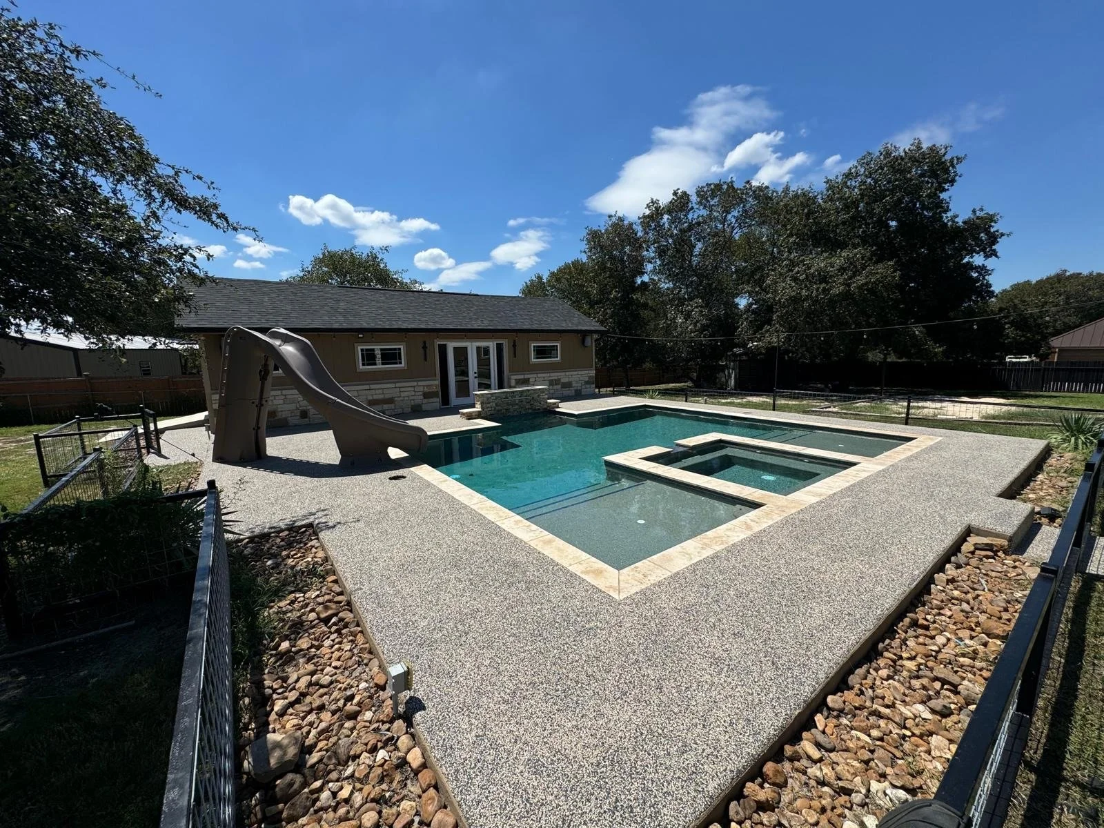 Backyard swimming pool with slide and hot tub, surrounded by gravel and fenced yard, under a blue sky with clouds.