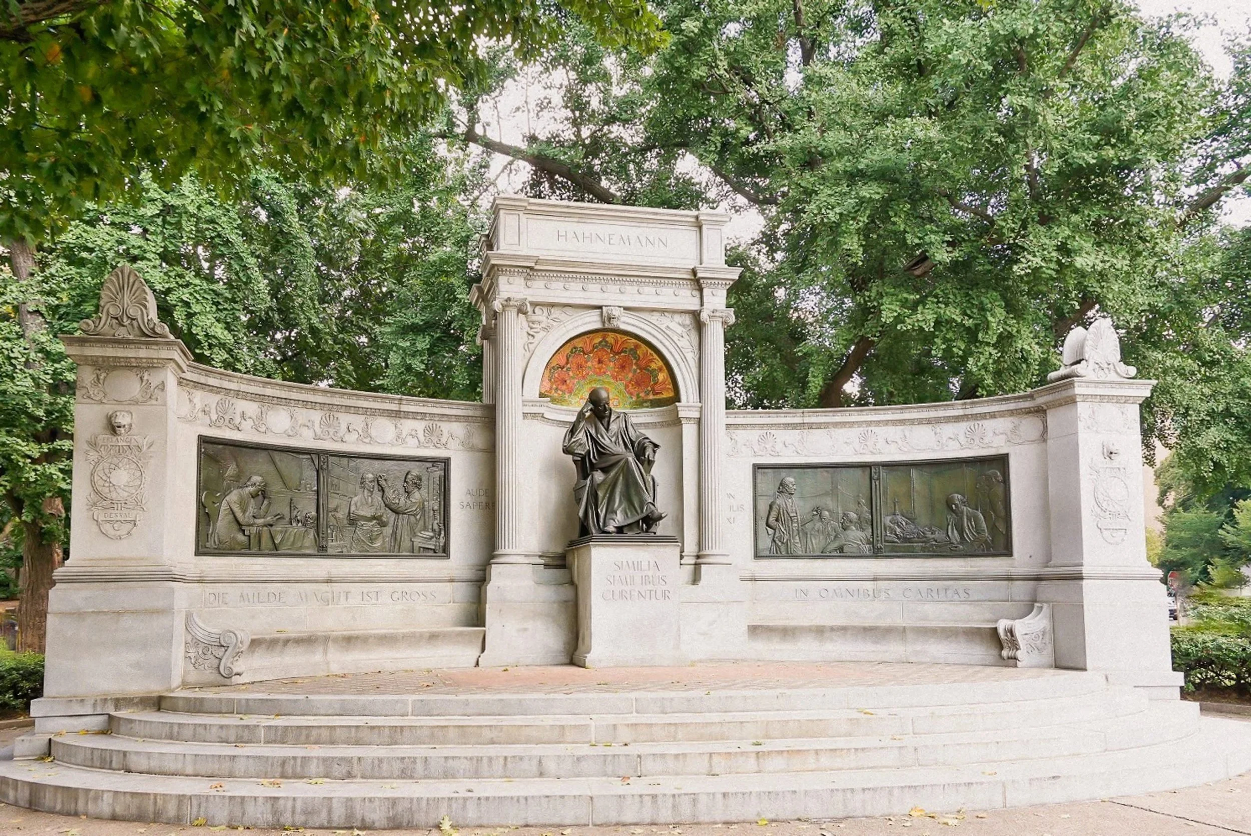 A monument with a statue of a seated man at the center, surrounded by decorative panels and carvings. The monument is set against green trees.