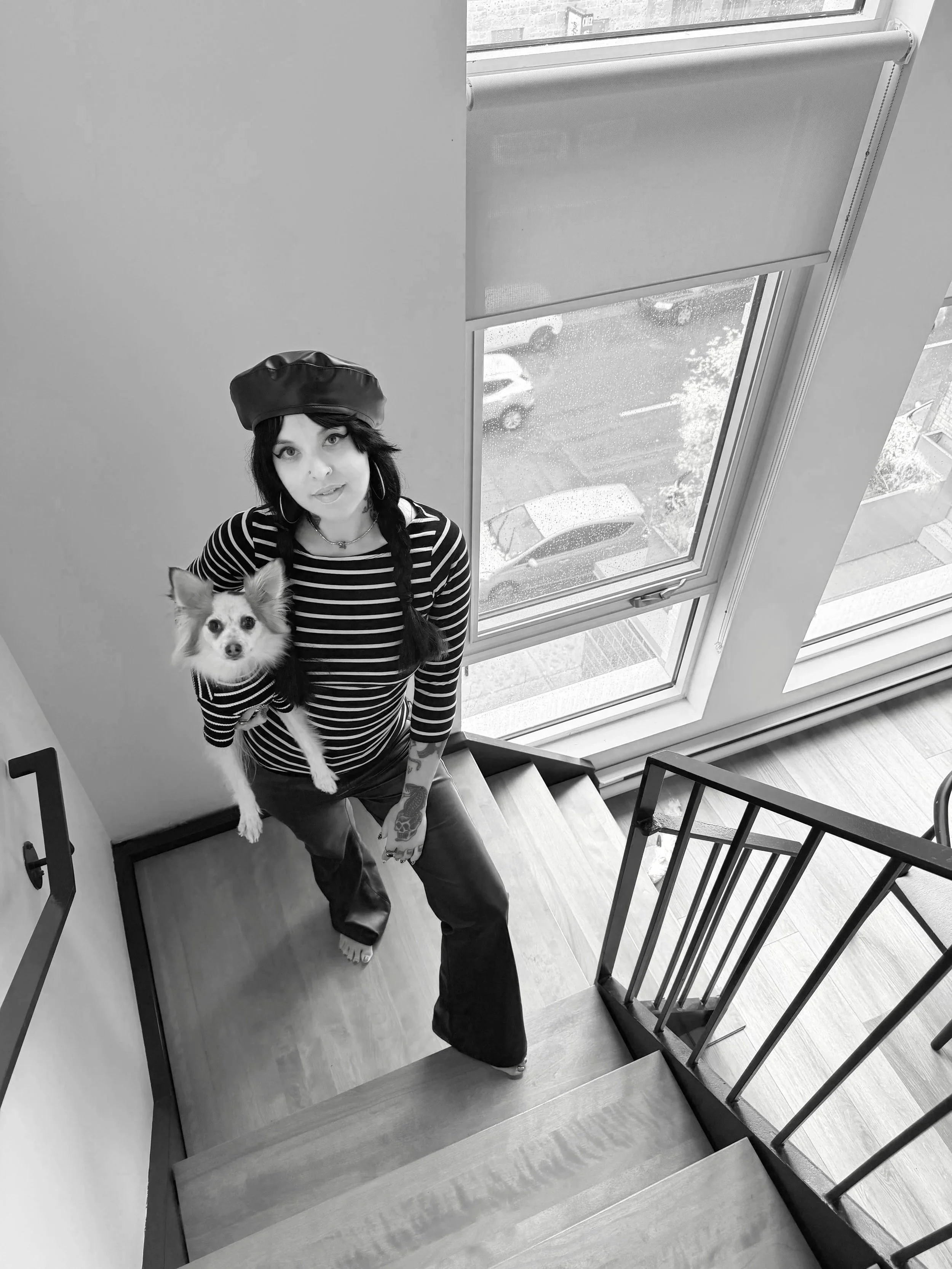 A woman with long dark hair in braids, wearing a beret and striped shirt, standing on a wooden staircase holding a small dog, with large windows showing a rainy street outside.