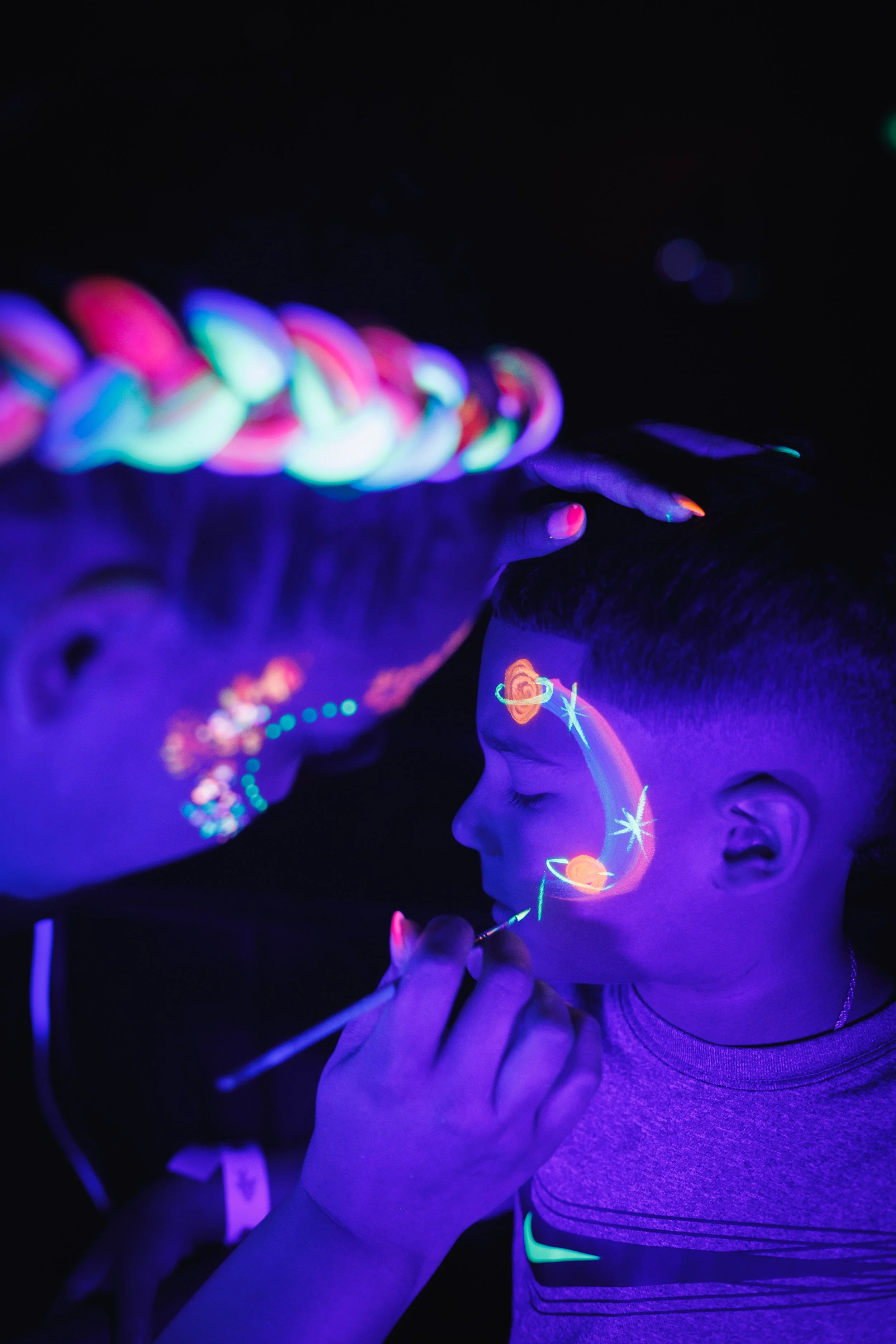 A child having their face painted with glow-in-the-dark colors at a dark venue, with the face painter also illuminated by black light.