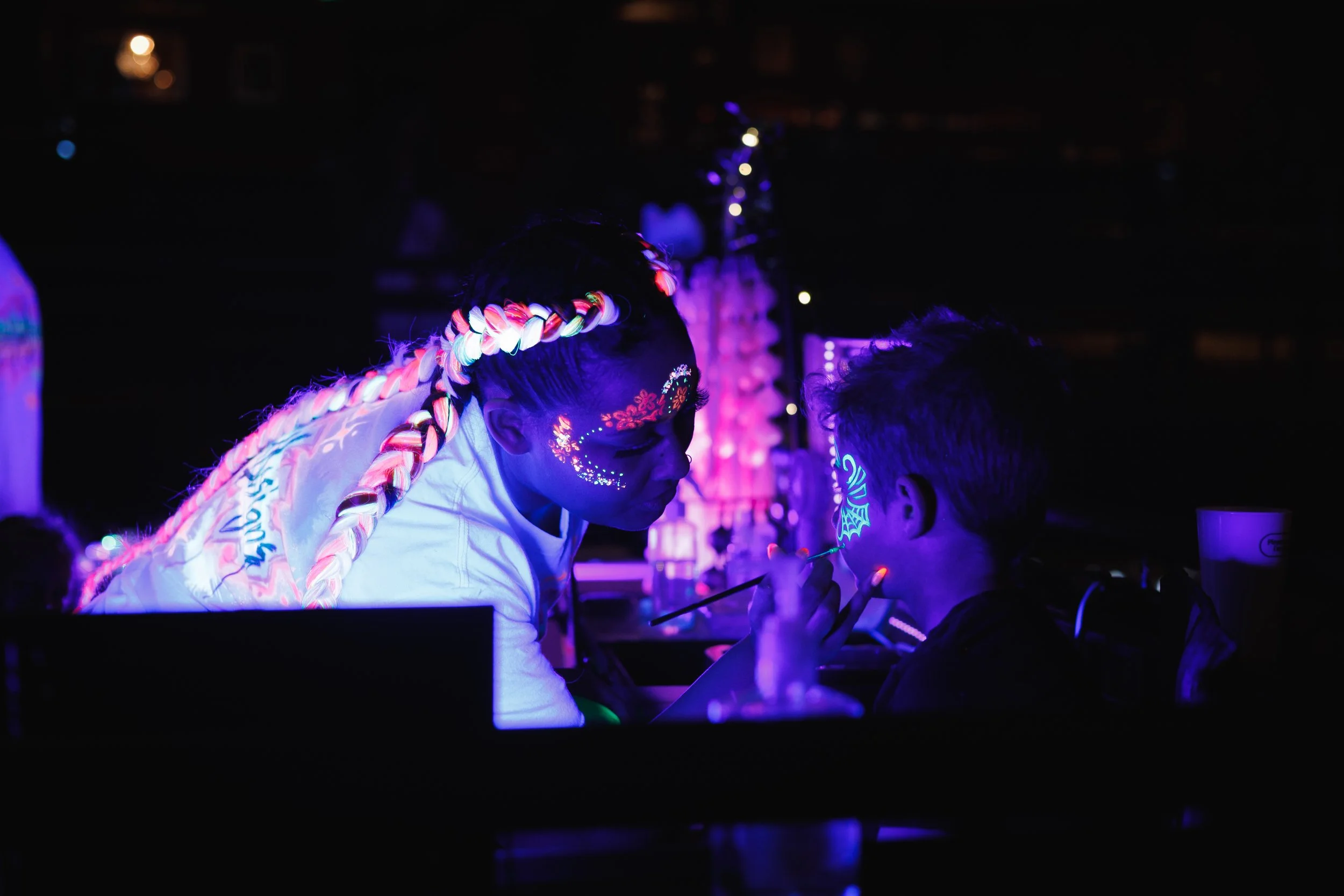 Two people with neon face paint and accessories exchanging face paint in a dark environment illuminated by blacklight, with colorful light decorations in the background.