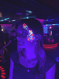 A woman at a nightclub with face painted in vibrant, colorful floral designs, illuminated by blue and purple lights.
