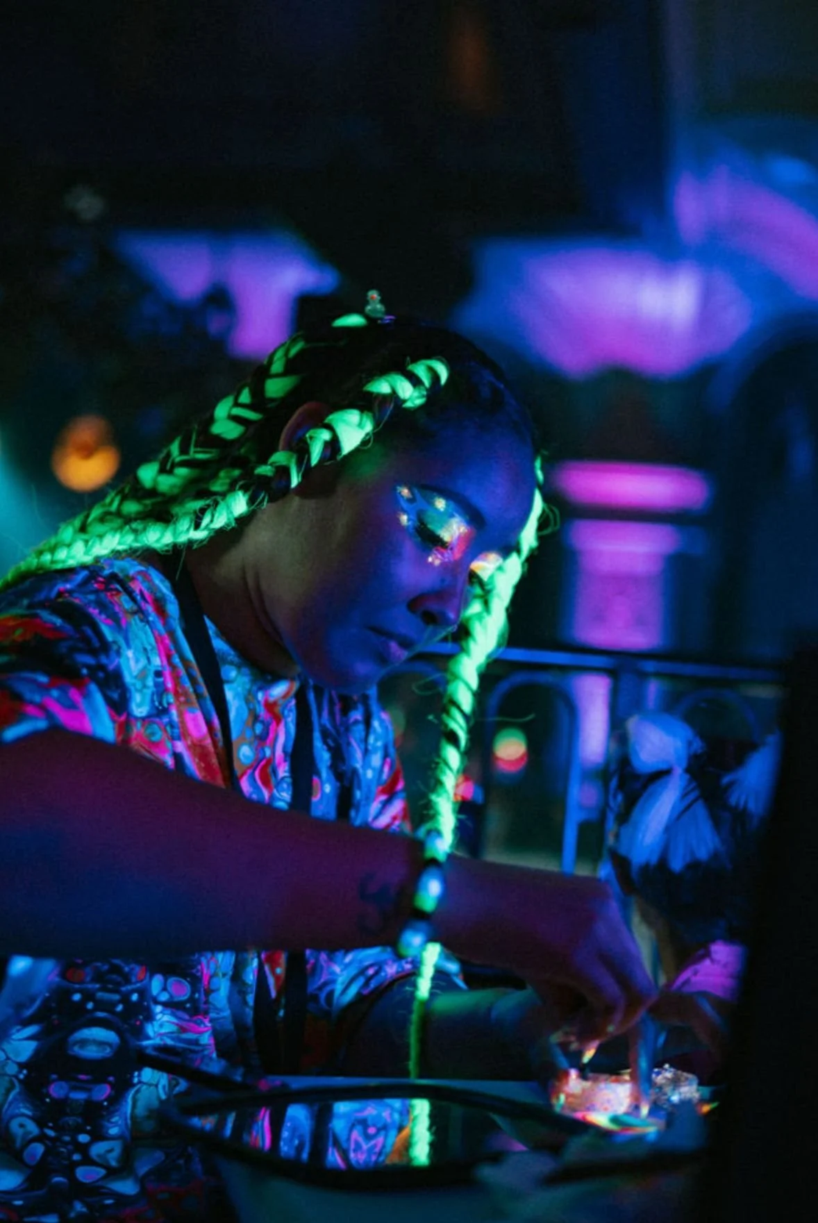 A woman with neon-colored braided hair, facial glitter, and vibrant clothing, working as a DJ in a dimly lit setting illuminated by blacklight glow.