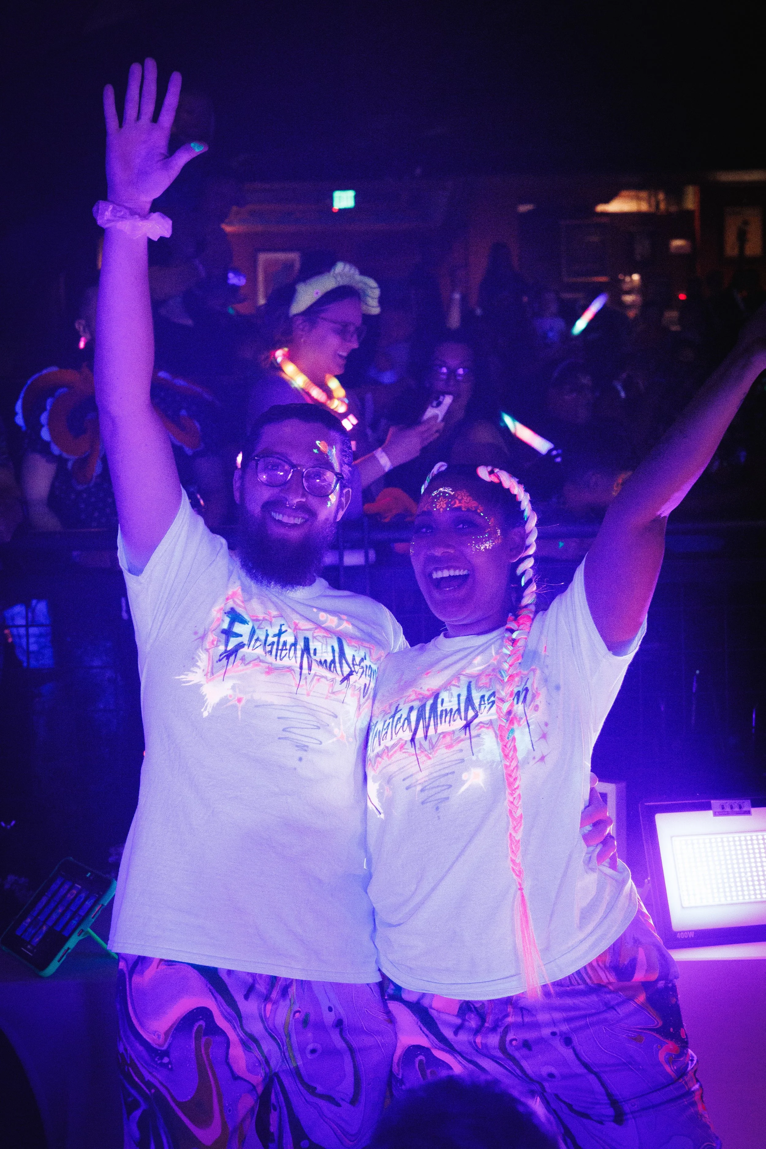 Two smiling people with glitter on their faces, wearing matching white T-shirts and psychedelic pants, celebrating with their arms raised at a fluorescent or glow-in-the-dark party.