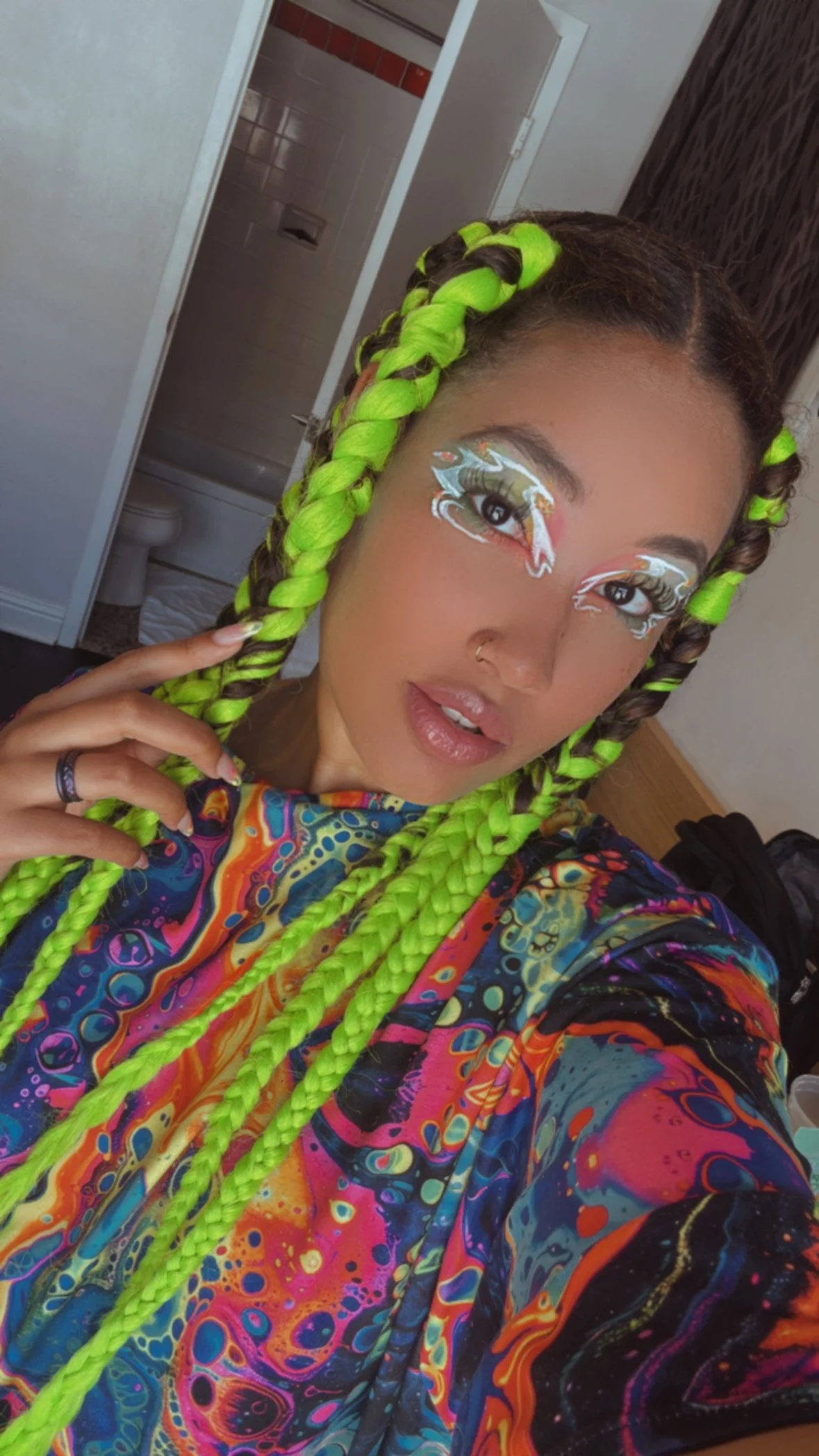 A woman with bright green braided hair and detailed fantasy-style eye makeup poses for a selfie in a room with a bathroom in the background.