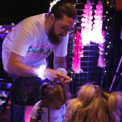 A man with glasses and a beard is applying face paint or makeup to a young girl with braided hair at a colorful event or festival, with pink and purple decorations in the background.