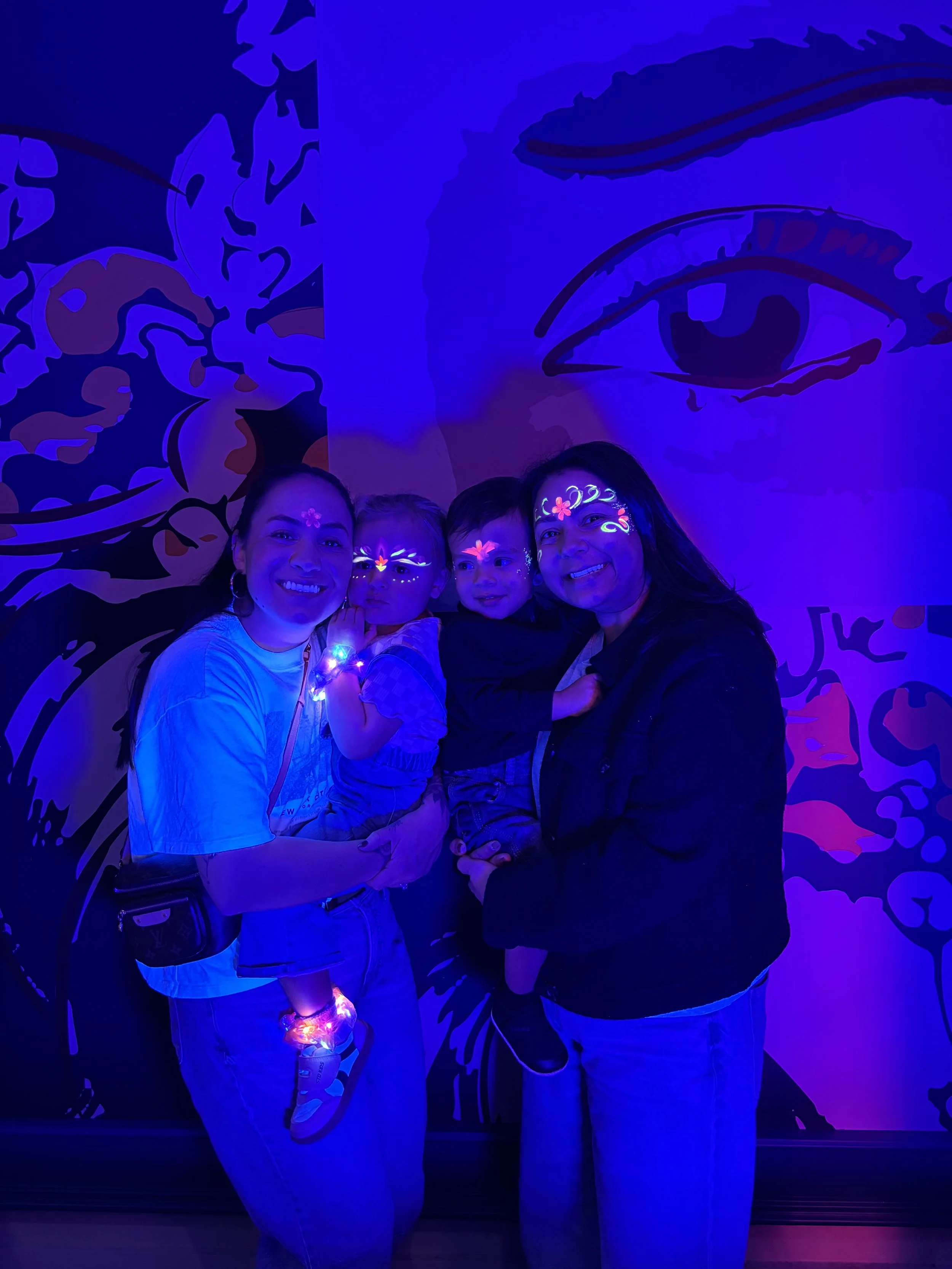 Group of four people, two women and two children, under blacklight with face paint and glowing accessories, against a wall with a large artistic eye and abstract pattern.