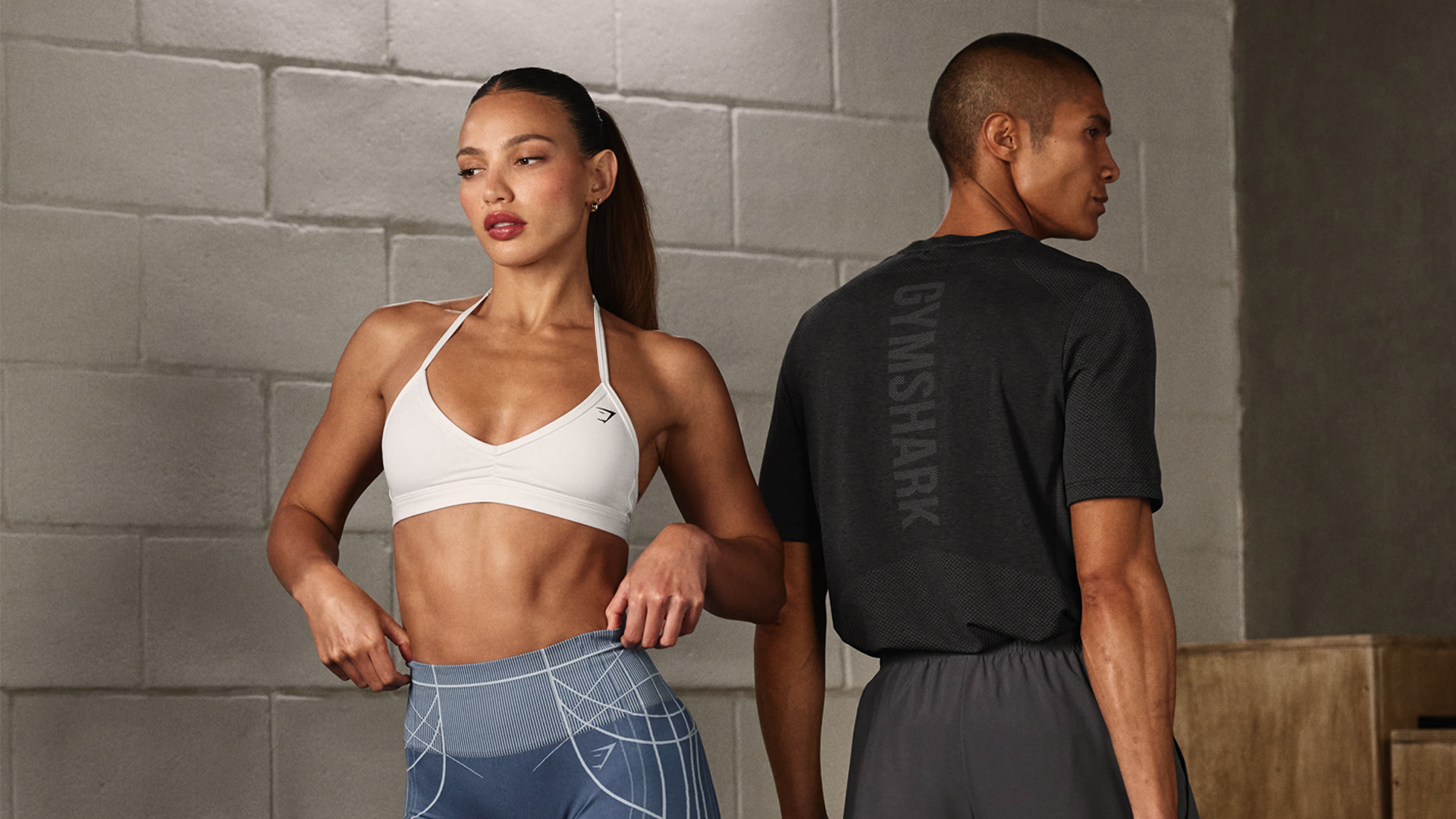 A woman in a white sports bra and patterned workout leggings standing with her hand on her hip, and a man in a black athletic shirt with 'GYMSHARK' written on the back and gray shorts, all in a gym setting.