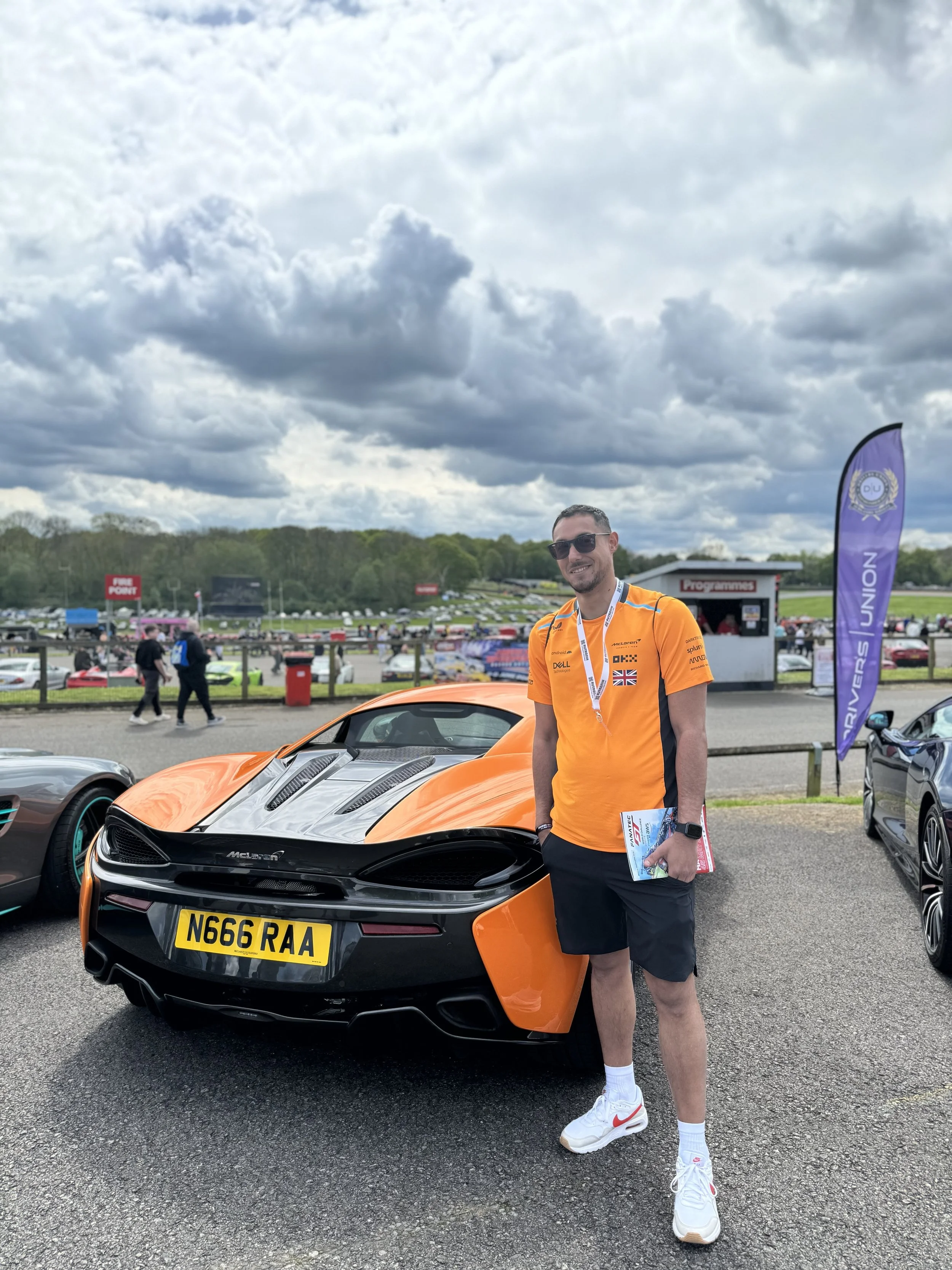Young man wearing sunglasses, an orange racing team shirt, and shorts standing next to a black and orange McLaren sports car at a racetrack event, with other cars and people in the background under a cloudy sky.
