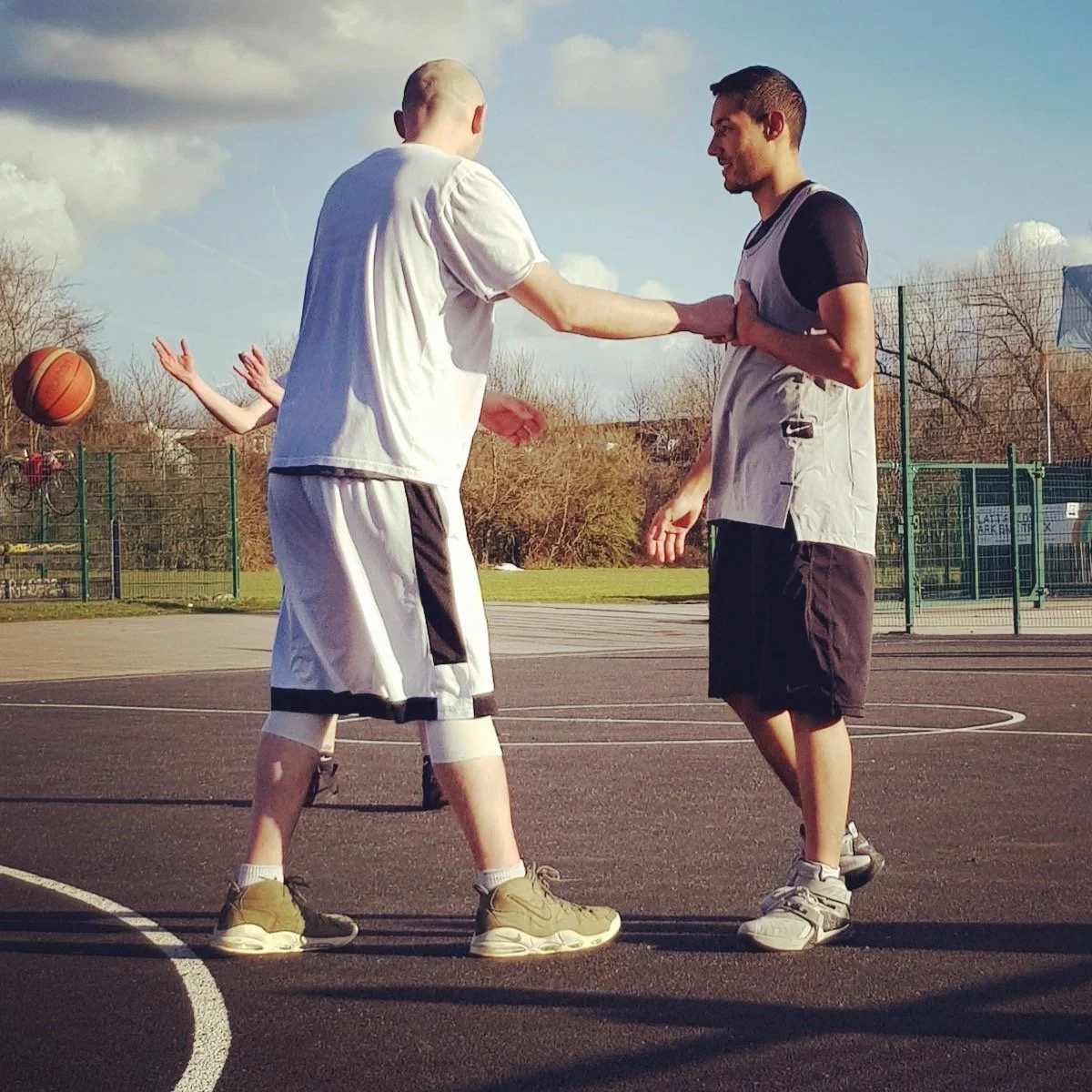 Two young men playing basketball on an outdoor court, one is reaching out to shake hands with the other, with a third person in the background. The sky is partly cloudy and trees are in the background.