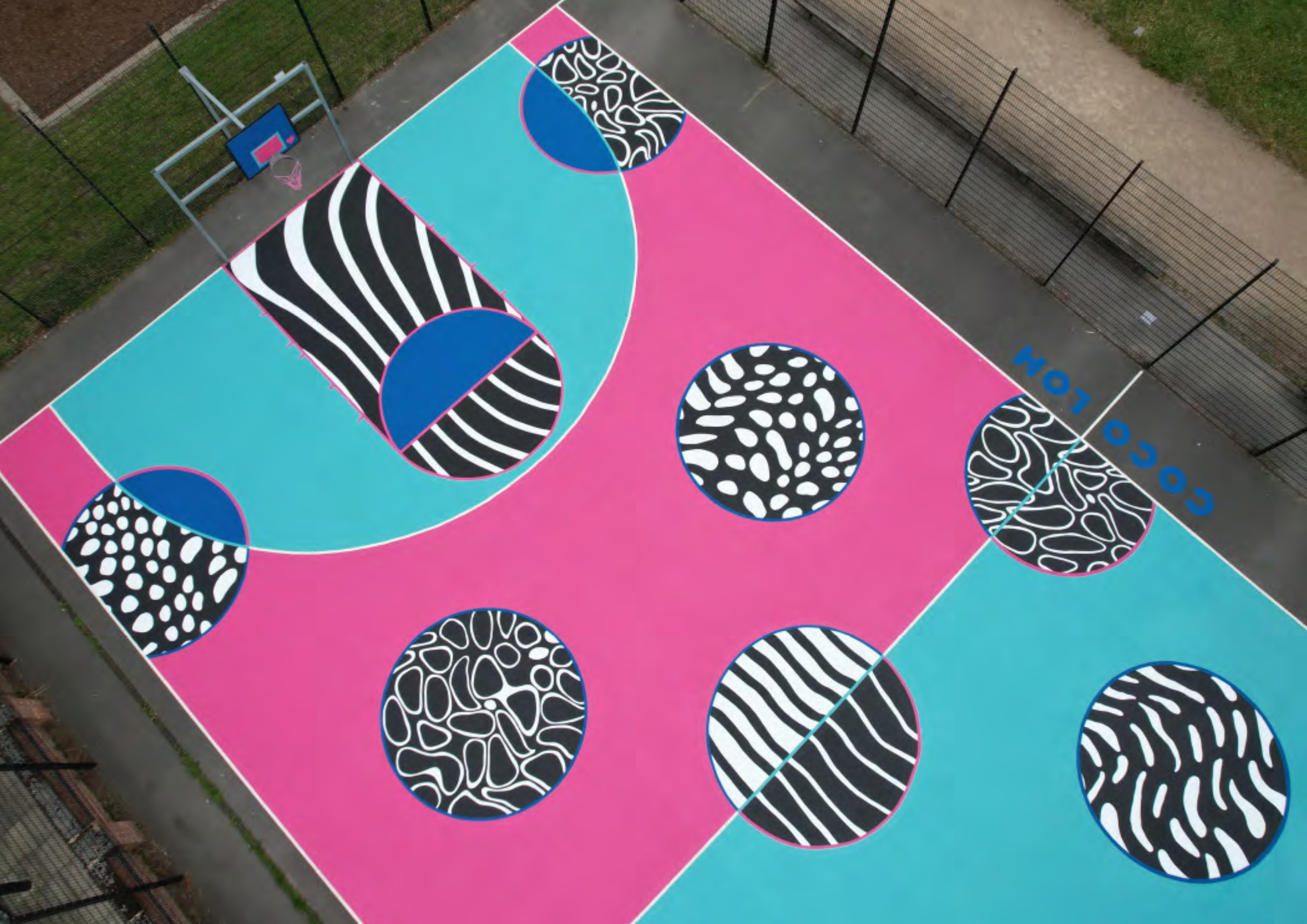 A colorful outdoor basketball court with a pink and blue surface featuring abstract black and white patterns inside circles. The court has a basketball hoop and backboard at one end and is enclosed by a black fence.