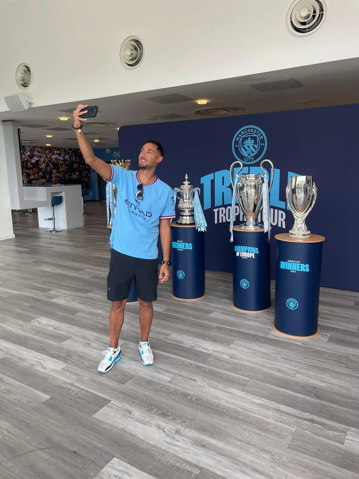 A man in a blue Manchester City soccer jersey taking a selfie at the Manchester City trophies display, which includes multiple trophies, in a room with a wooden floor and a dark blue backdrop.
