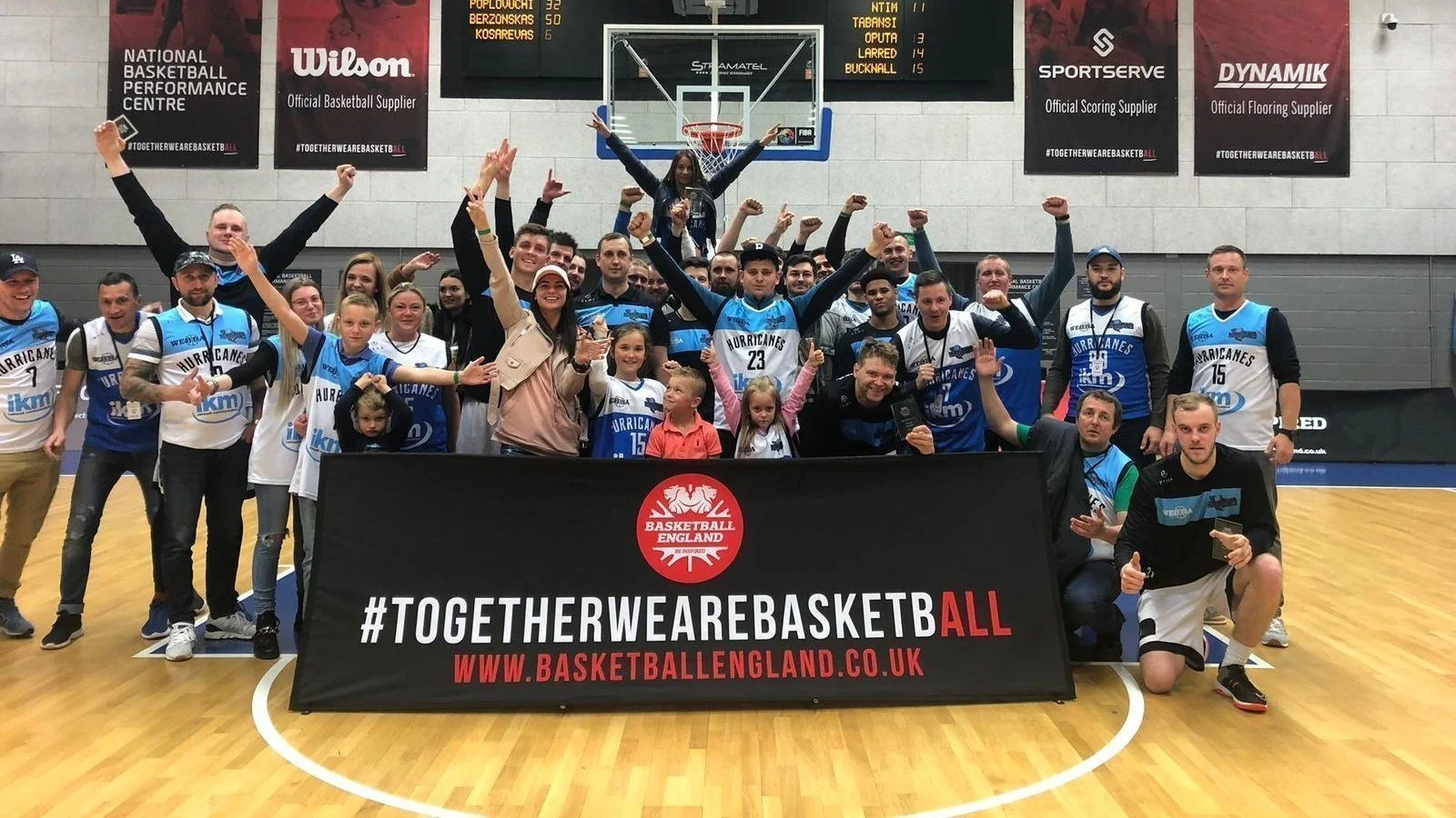 A group of people, including basketball players and children, posing on an indoor basketball court with a banner that reads #TogetherWeAreBasketball and www.basketballengland.co.uk, celebrating their participation in basketball.