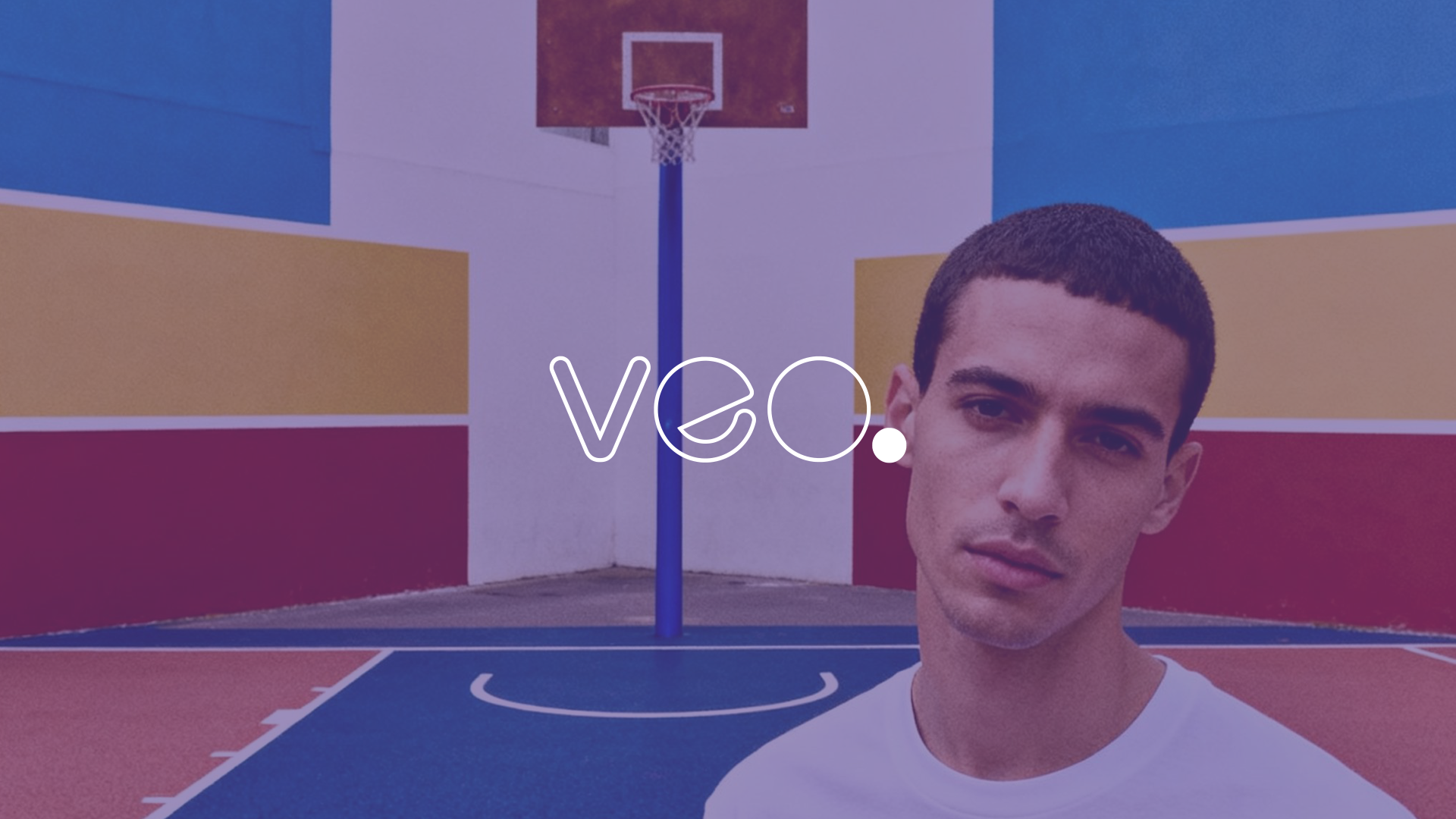 A man with short dark hair standing in front of a colorful outdoor basketball court with red, yellow, blue, and white walls, and a basketball hoop in the background.