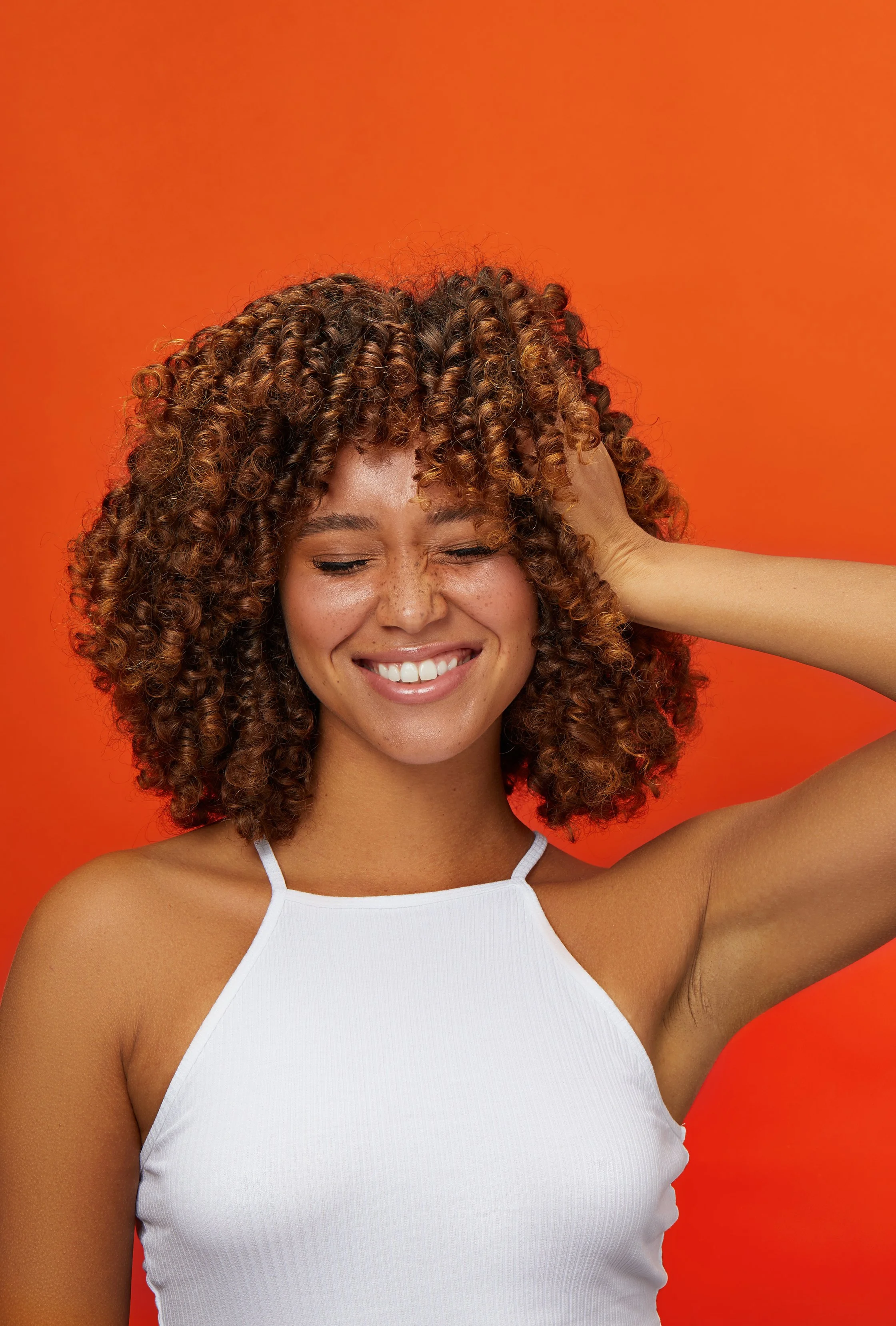 Young woman smiling with curly hair, wearing a white tank top, against an orange background.