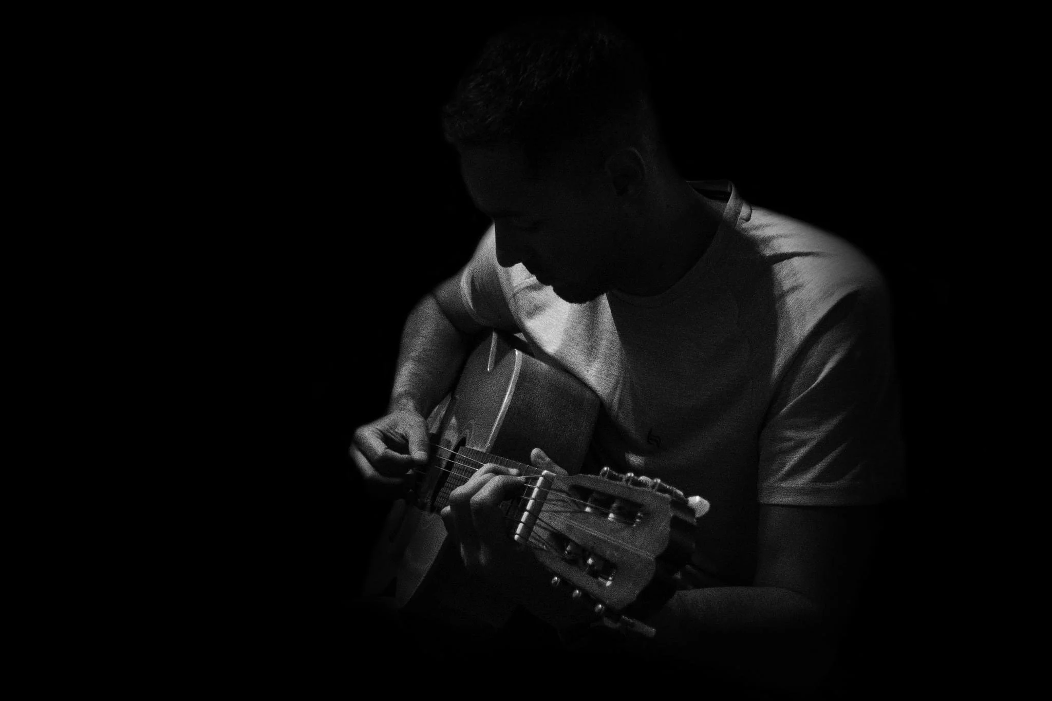 Black and white photo of a man playing an acoustic guitar, with his face in shadow, against a dark background.