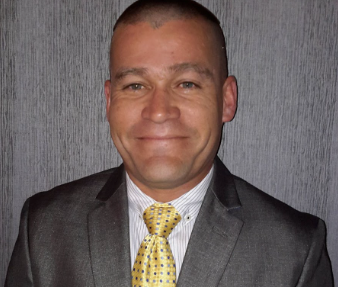 A man in a gray suit, white shirt, and yellow tie smiling in front of a gray textured background.