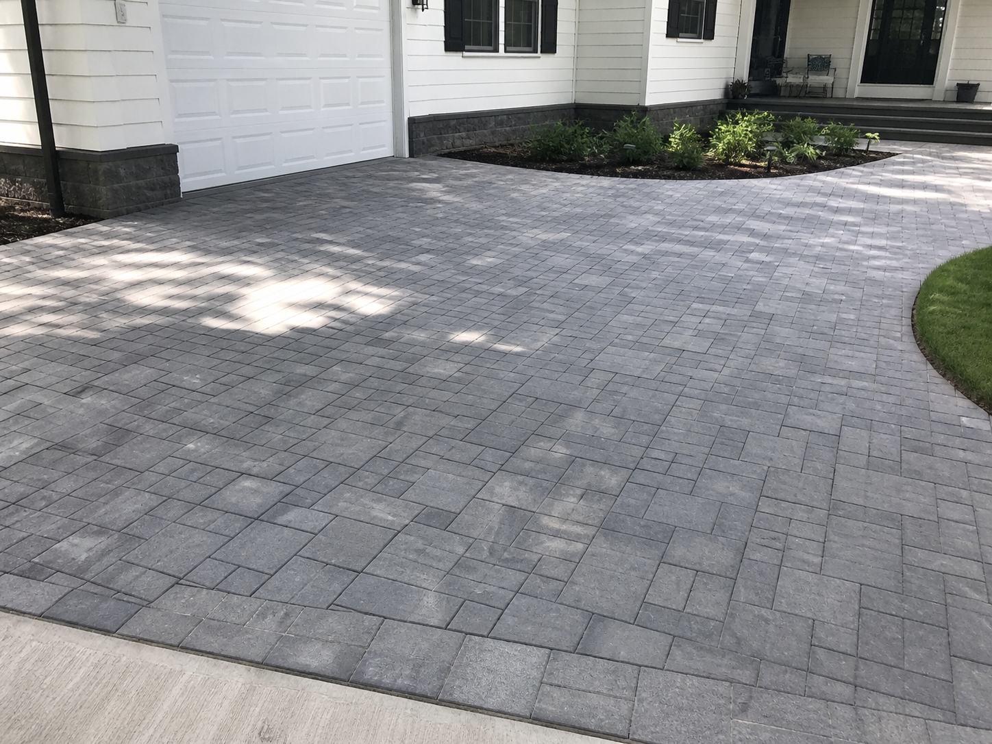 Gray stone paver driveway leading to a white garage door and a house with a small flower bed and steps.