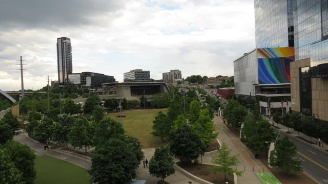 Cityscape with modern buildings, a park with trees and walking paths, and a cloudy sky.