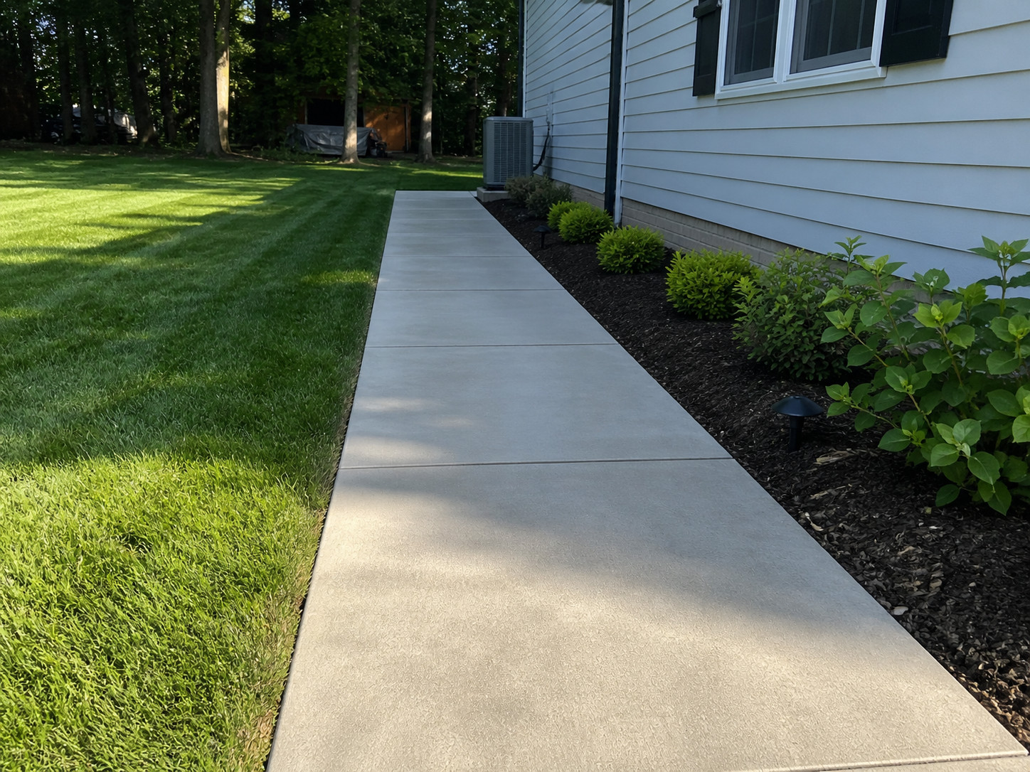 A sidewalk running alongside a house with green shrubs and small landscape lights in a backyard. The grass yard is on the left, and there are trees and a shed in the background.
