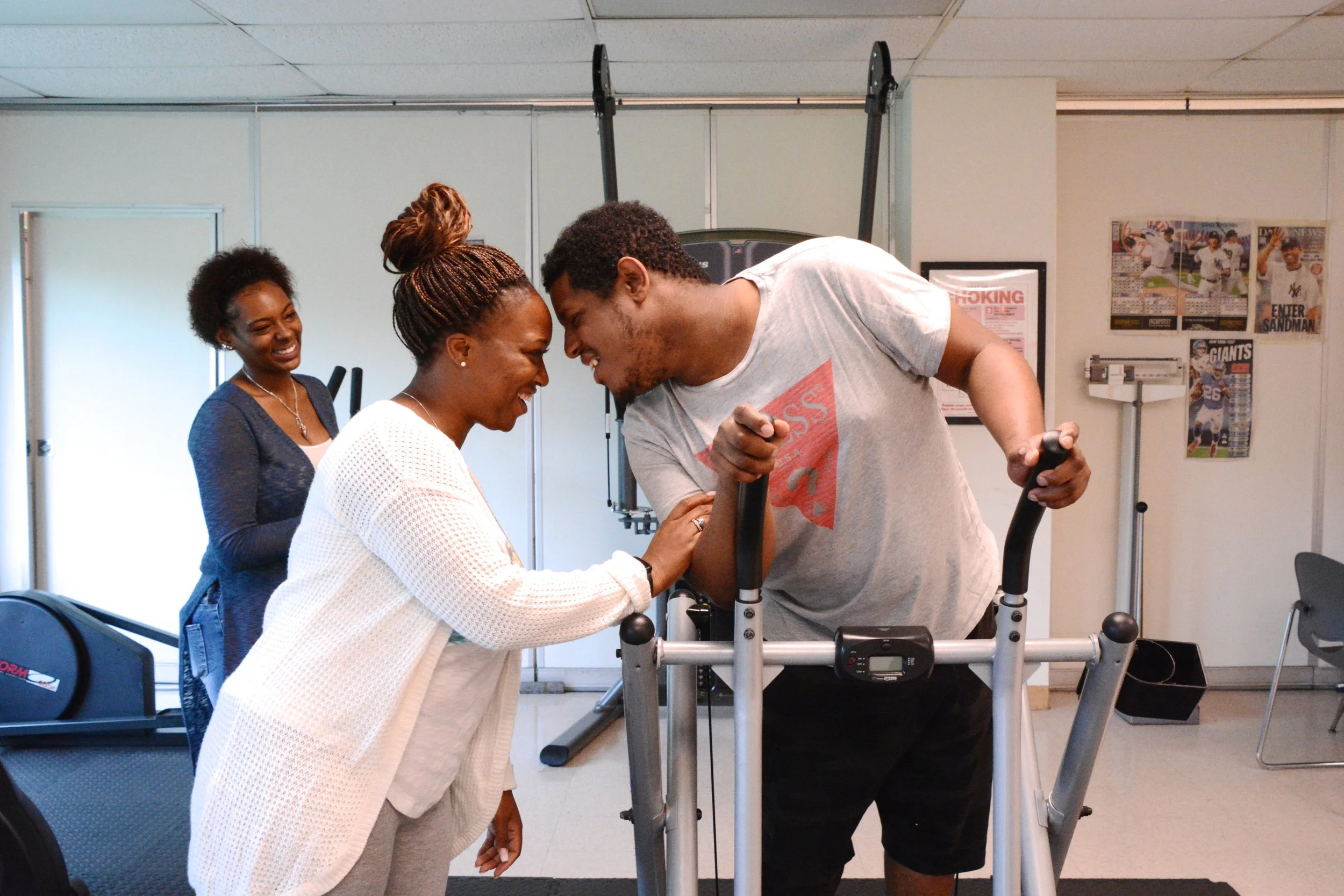 A man using a walker shares a victory moment with a woman at a rehabilitation center, while another woman watches smiling.