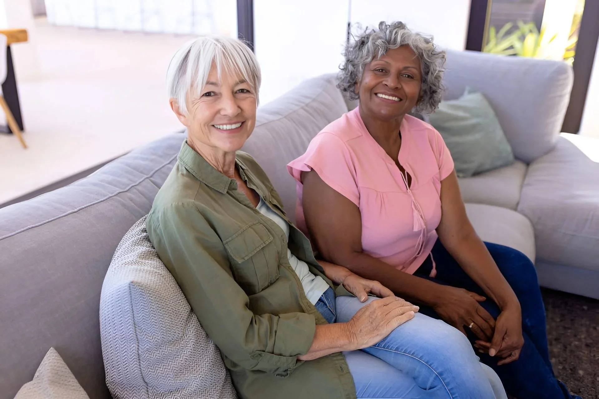 Two smiling older women sitting on a light-colored sofa in a bright, modern living room.