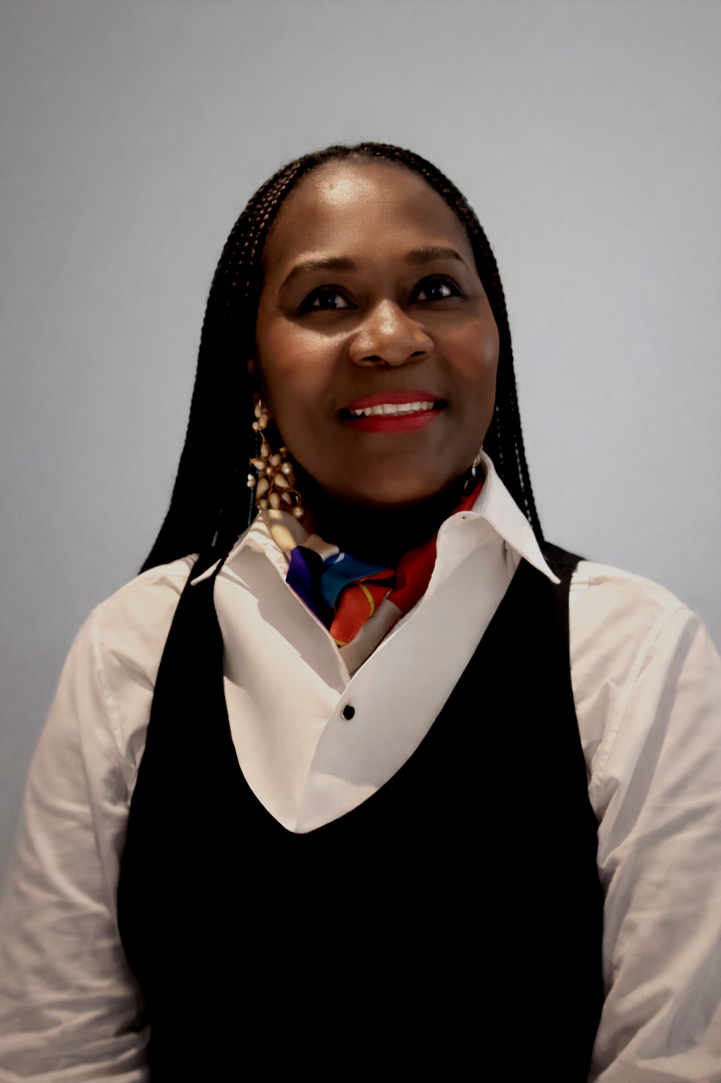 A woman with dark braided hair, wearing a white collared shirt, black vest, colorful scarf, and large earrings, smiling and looking to the side against a plain gray background.