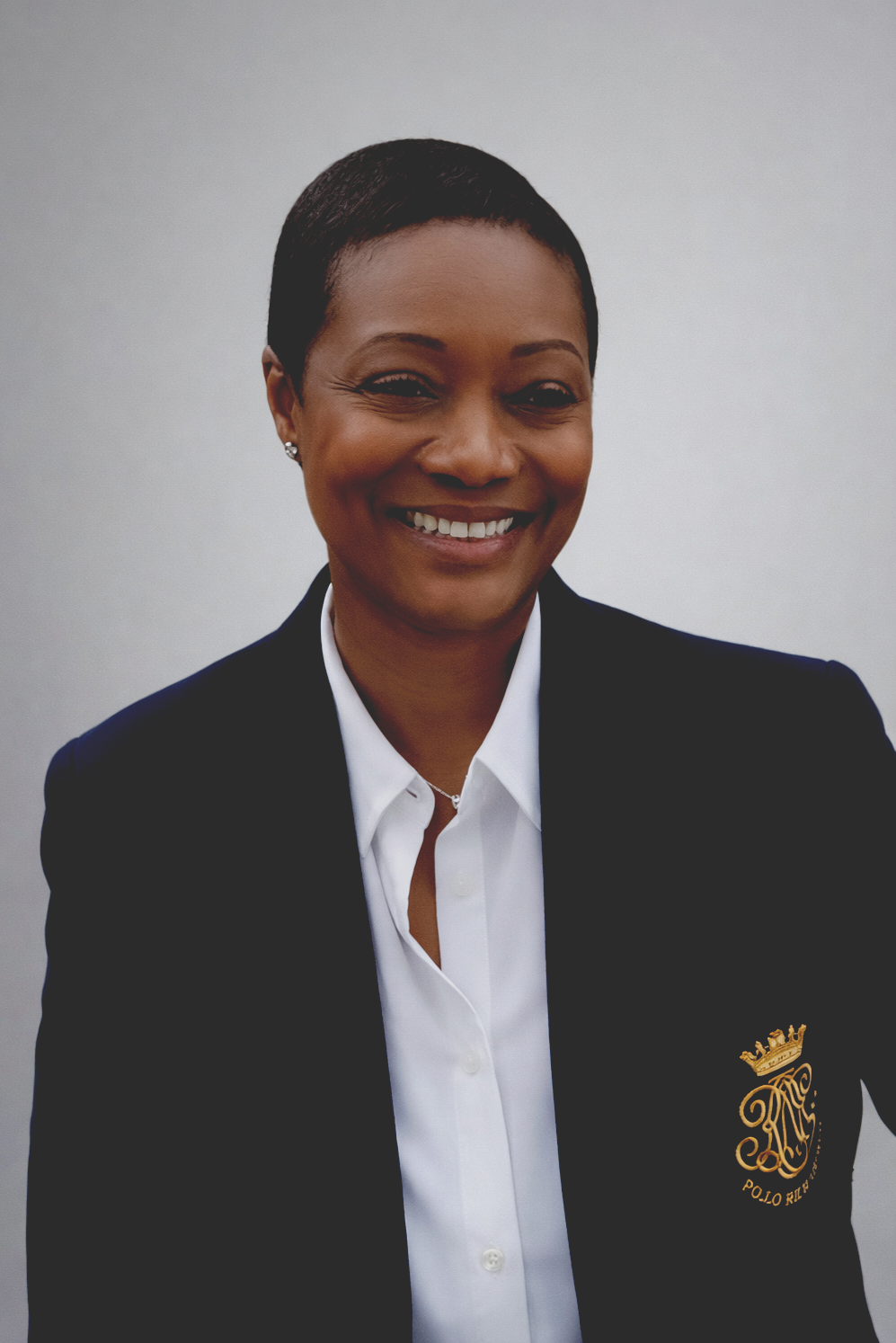 Professional woman with short haircut smiling, wearing a black blazer with a gold embroidered crest and a white shirt, against a gray background.