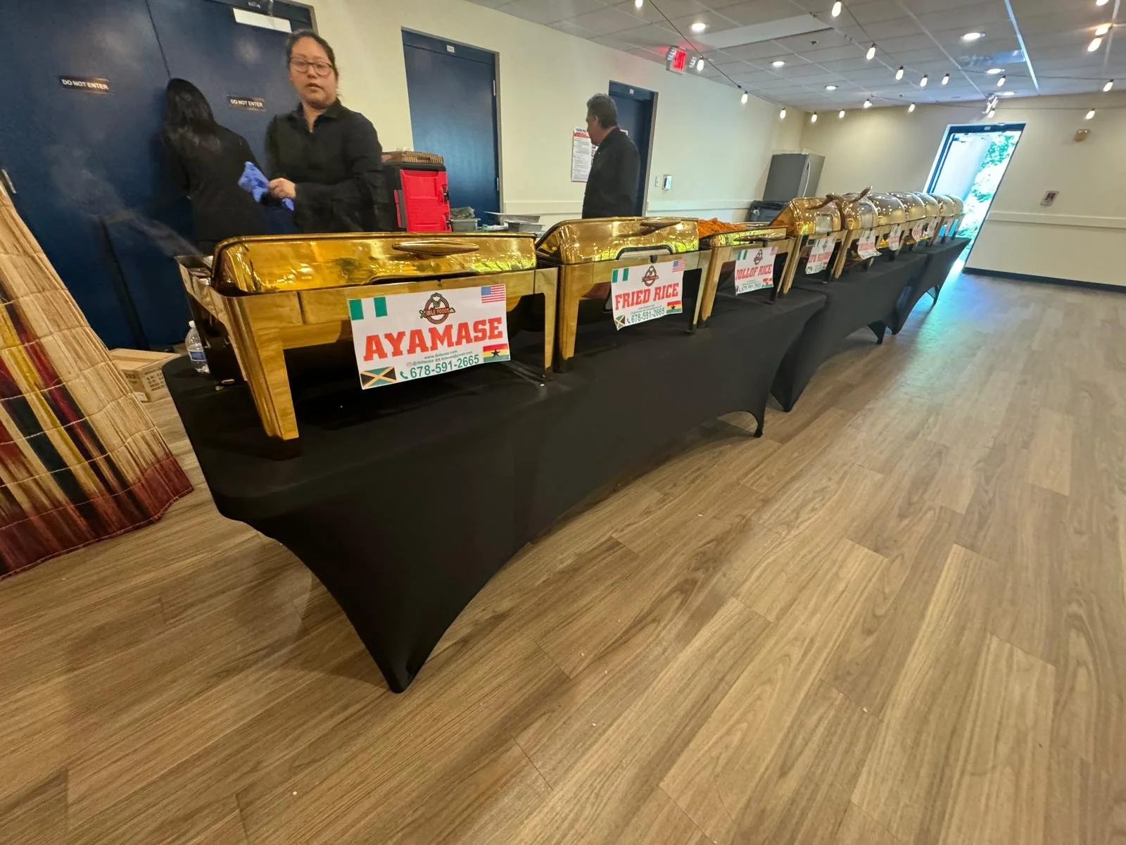 A buffet table with chafing dishes labeled for different dishes, including 'AYAMASE' and 'FRIED RICE,' in a dining area with people in the background.