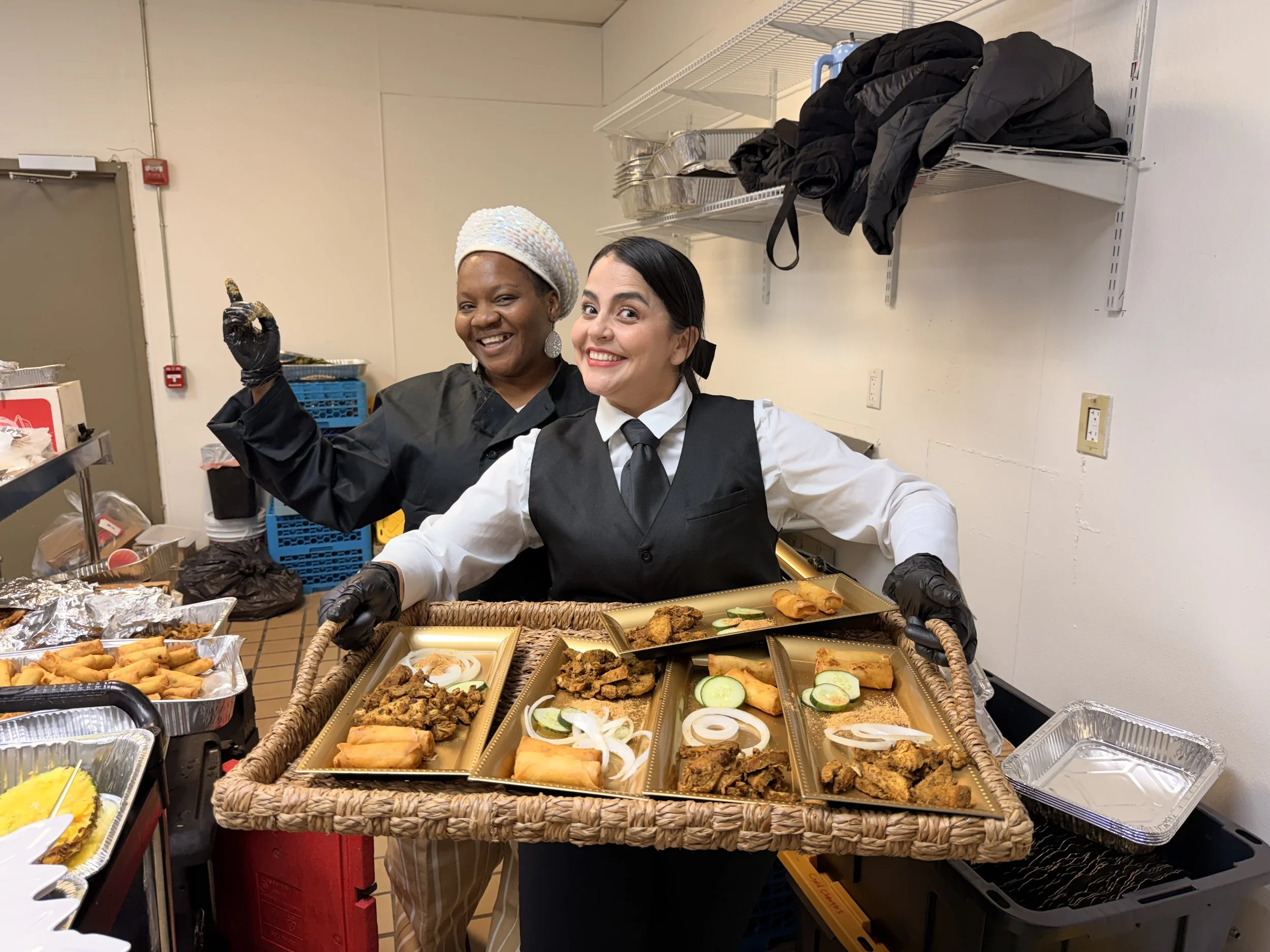 Two women in a kitchen smiling and holding a tray of assorted fried foods, vegetables, and spring rolls, with food preparation items around them.