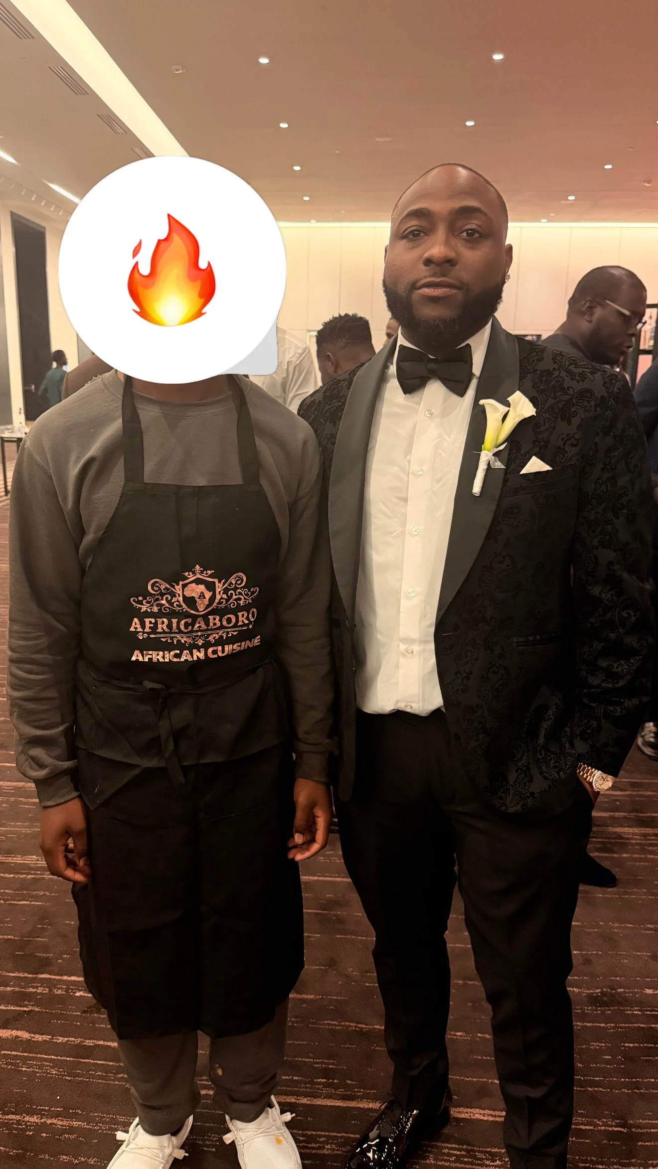 A man in a tuxedo standing next to a person with face covered by a fire emoji, in a formal event setting.