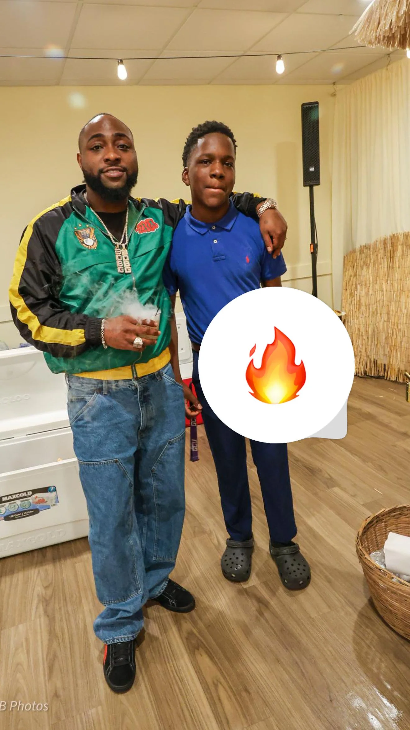 Two men standing indoors with one having an arm around the other's shoulder. One man is holding a smoking object. A speech bubble with a fire emoji covers part of the image.