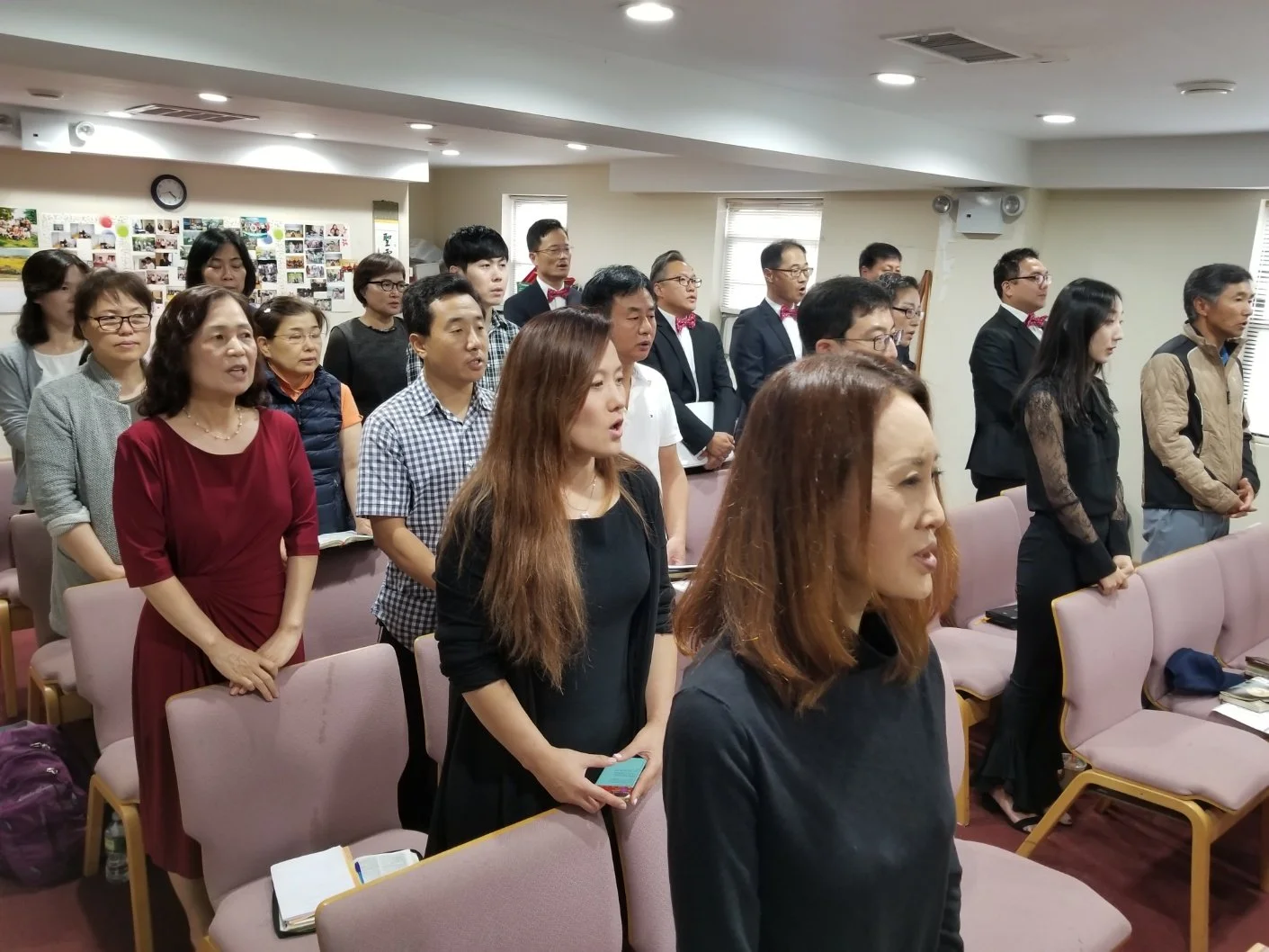 A group of diverse people standing in a room during a formal event or ceremony, facing forward with their eyes closed or looking attentively, with some holding notebooks or phones, and a wall with photos and a clock in the background.