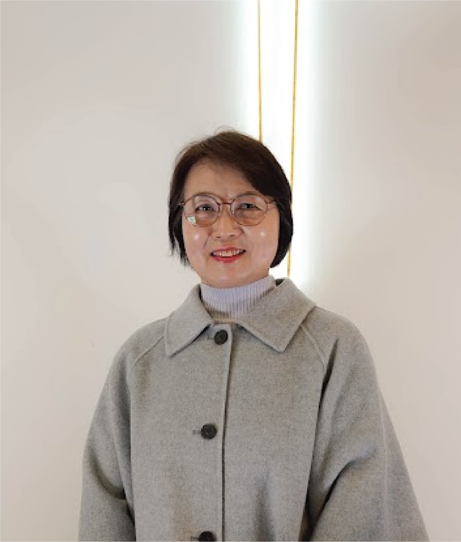 A woman with short dark hair, glasses, and light makeup smiling and wearing a light gray coat over a white turtleneck sweater, standing indoors against a plain white wall with vertical lighting.
