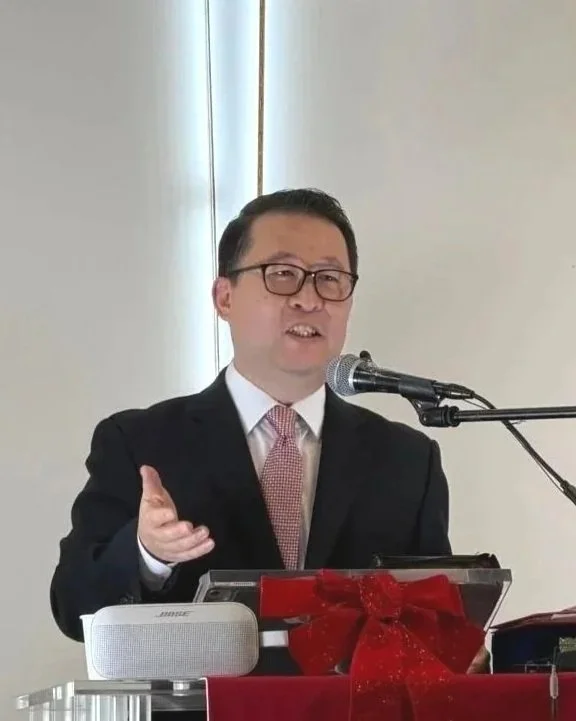 Man in a suit speaking into a microphone at a podium decorated with a red ribbon and bow.