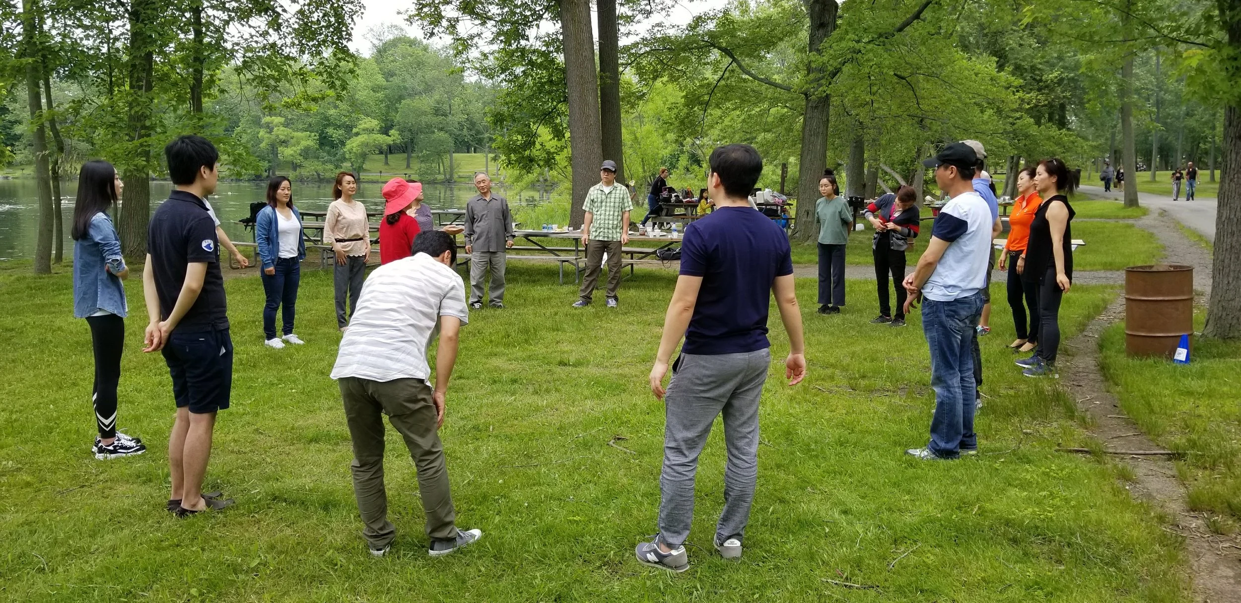 People gathered outdoors in a circle on a grassy area near a lake, participating in an activity or game, with trees and picnic tables in the background.