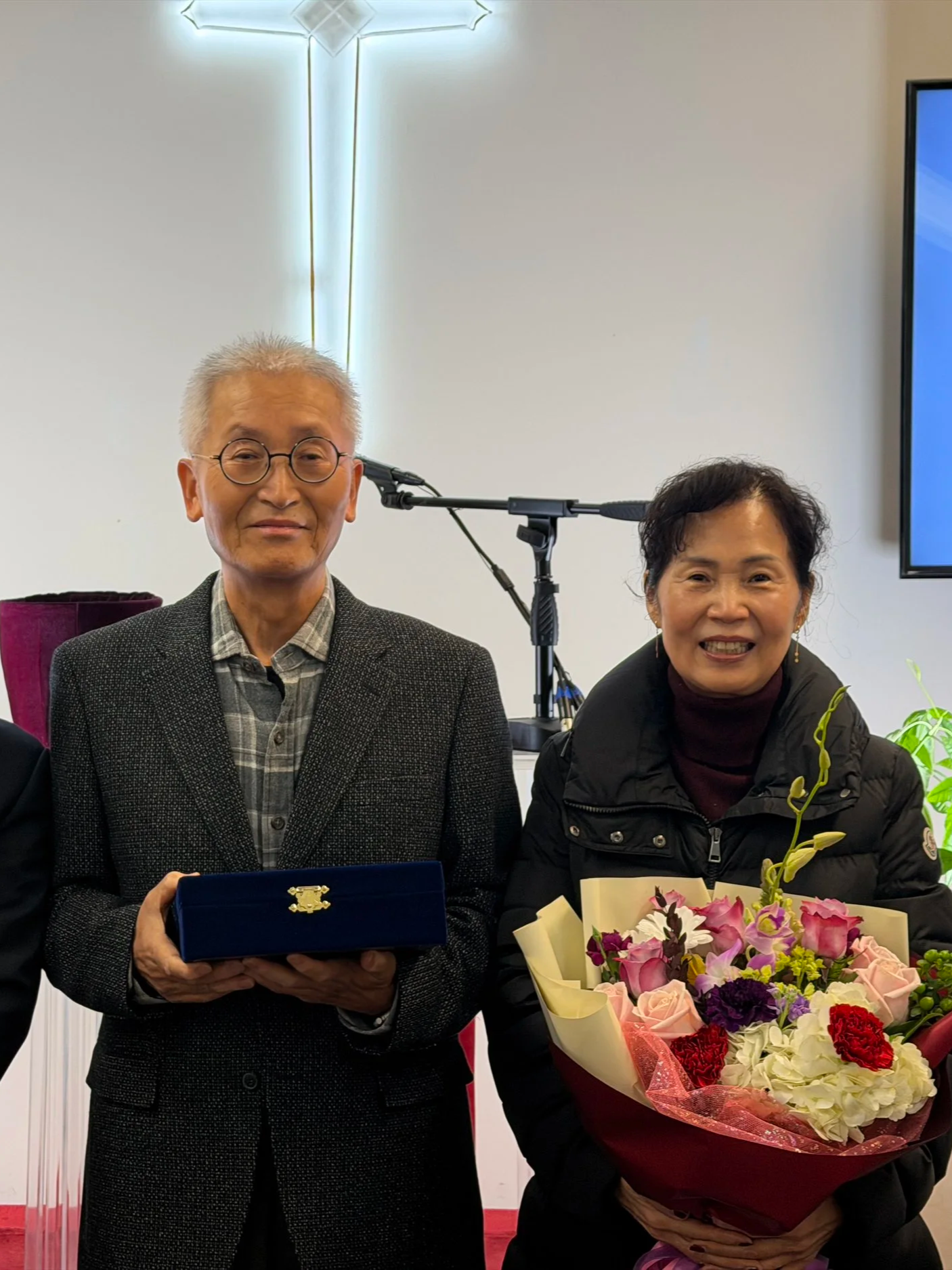 An elderly man with glasses wearing a suit and a woman sitting next to him holding a bouquet of flowers.