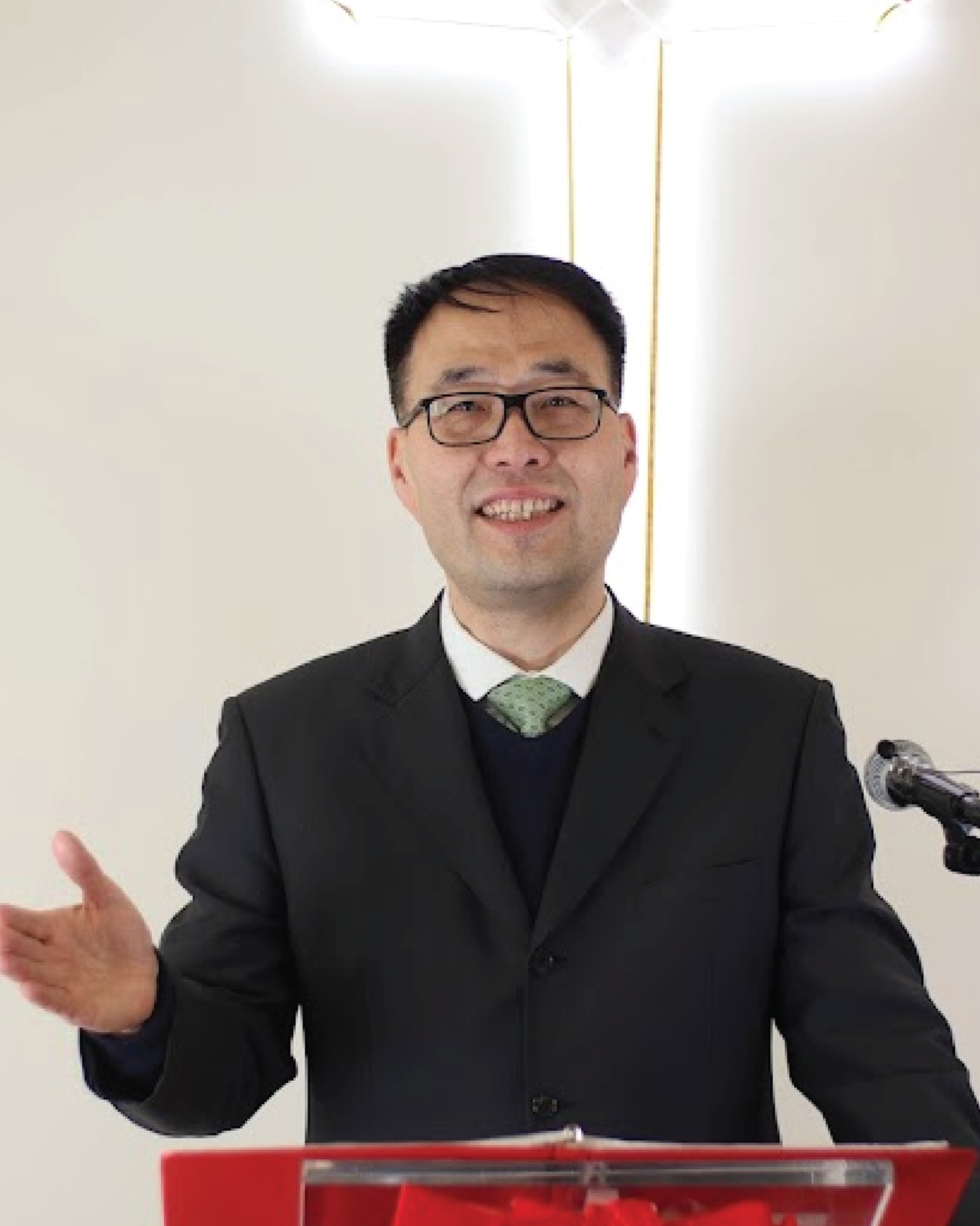 A man wearing glasses, a black suit, white shirt, and green tie is speaking at a podium.