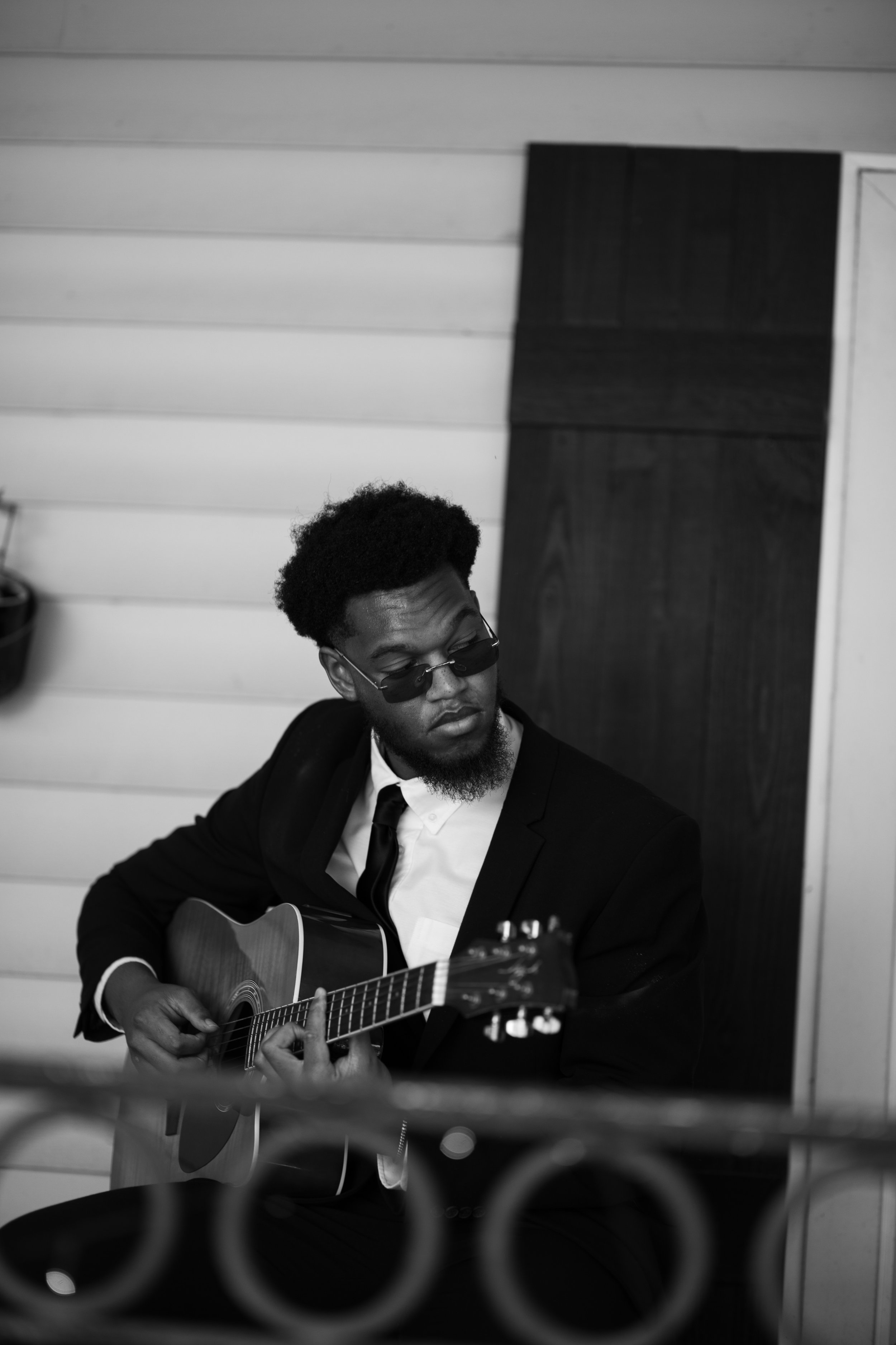 A man wearing sunglasses and a suit, playing an acoustic guitar.