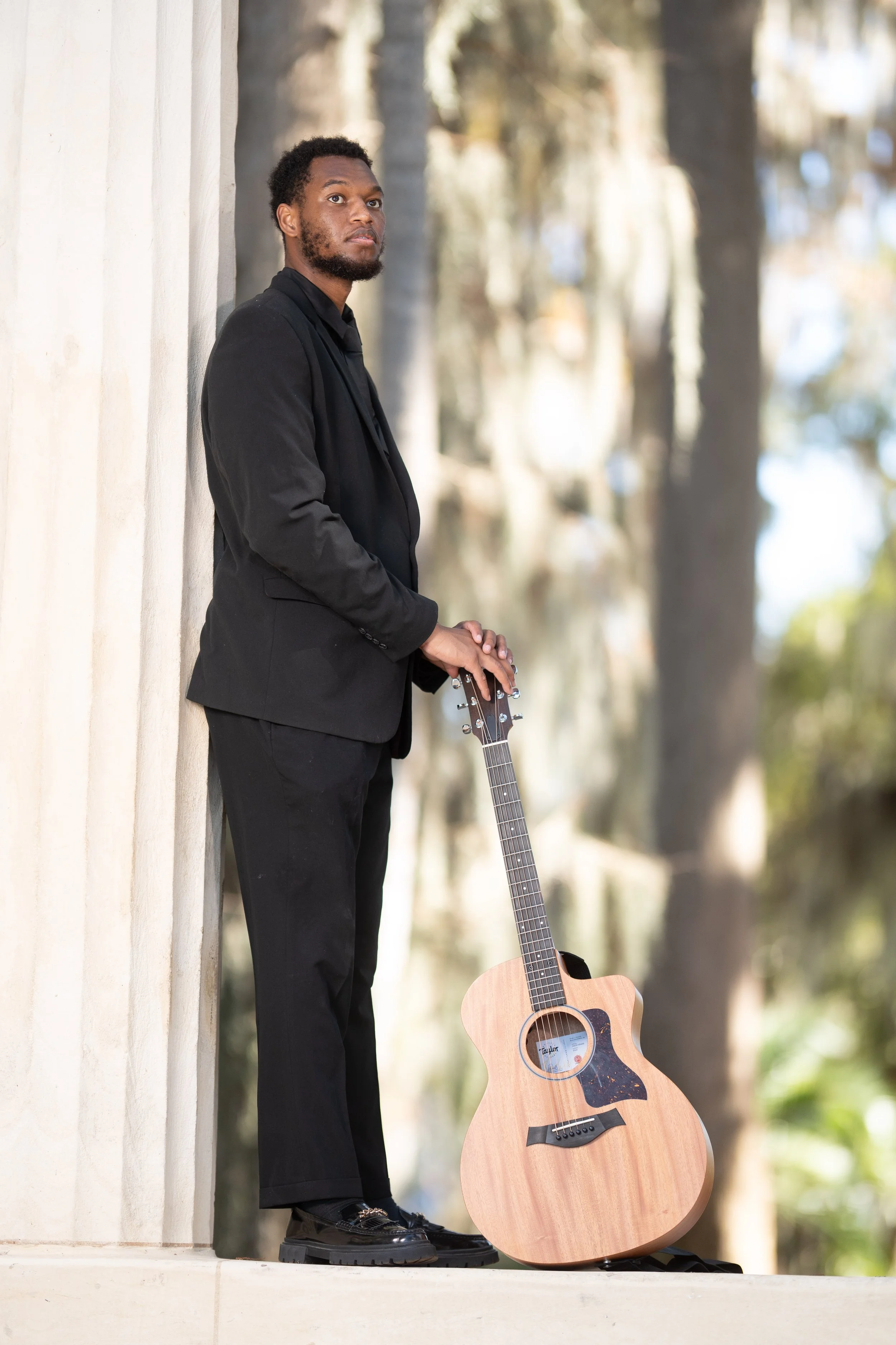A man in a black suit leaning against a beige wall, holding an acoustic guitar outdoors with trees in the background.