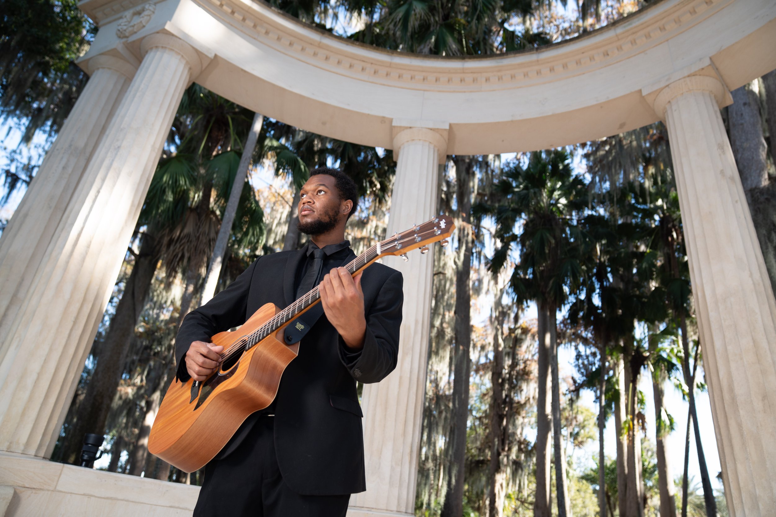 A man in a black suit playing an acoustic guitar under a neoclassical columned pavilion, with tall trees in the background.