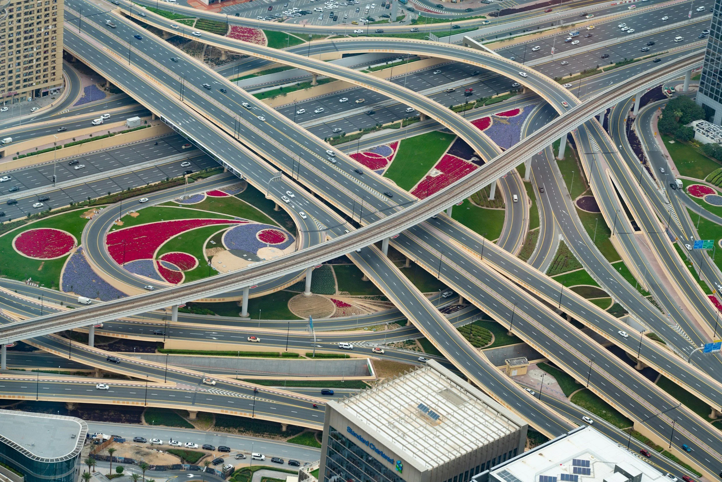 Aerial view of multiple overpasses, roads, and a landscaped area with colorful flower beds in geometric patterns in an urban setting.