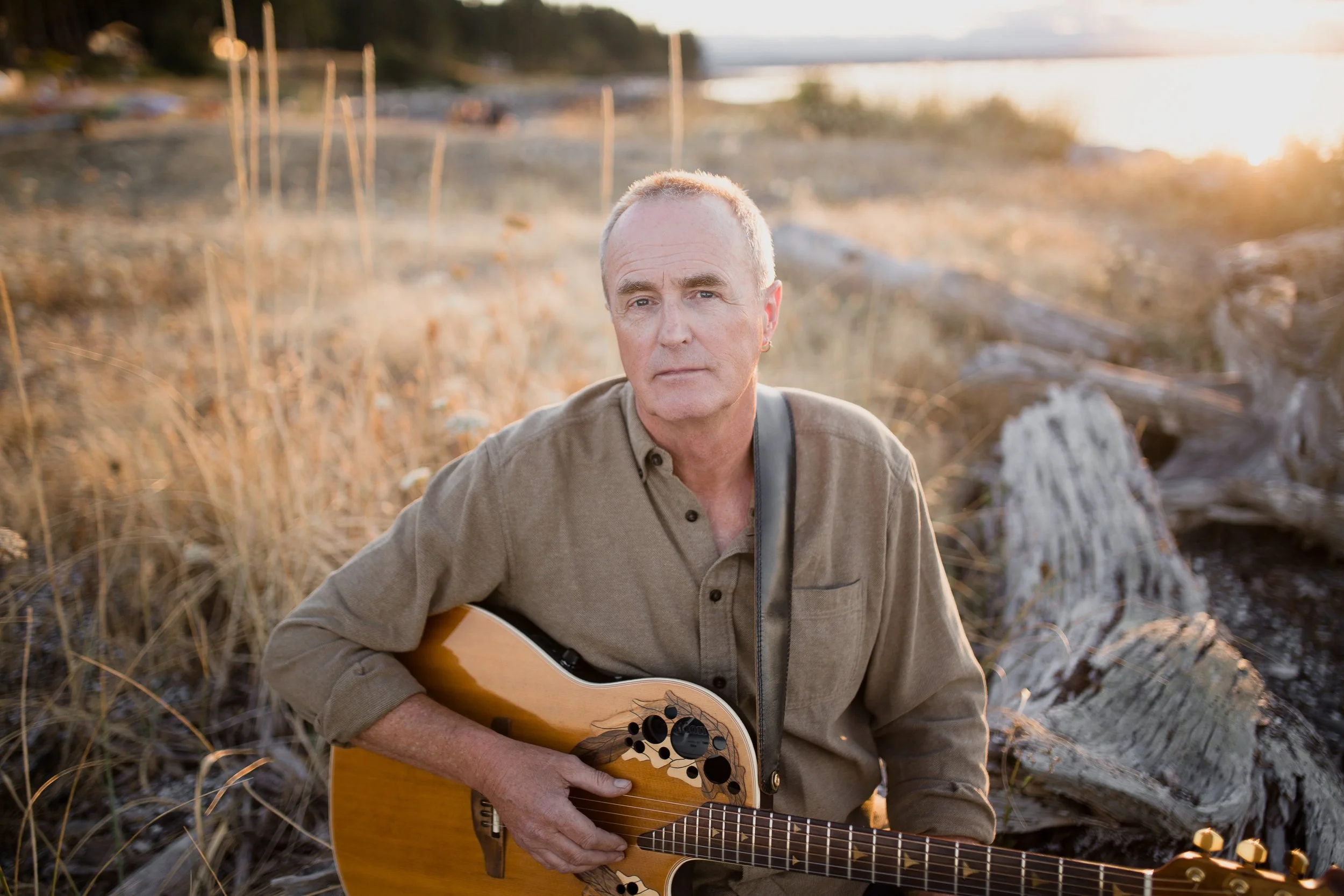 Terry Boyle, Vancouver Island singer songwriter, playing an acoustic guitar outdoors during sunset, surrounded by dry grass and fallen logs on Hornby Island.