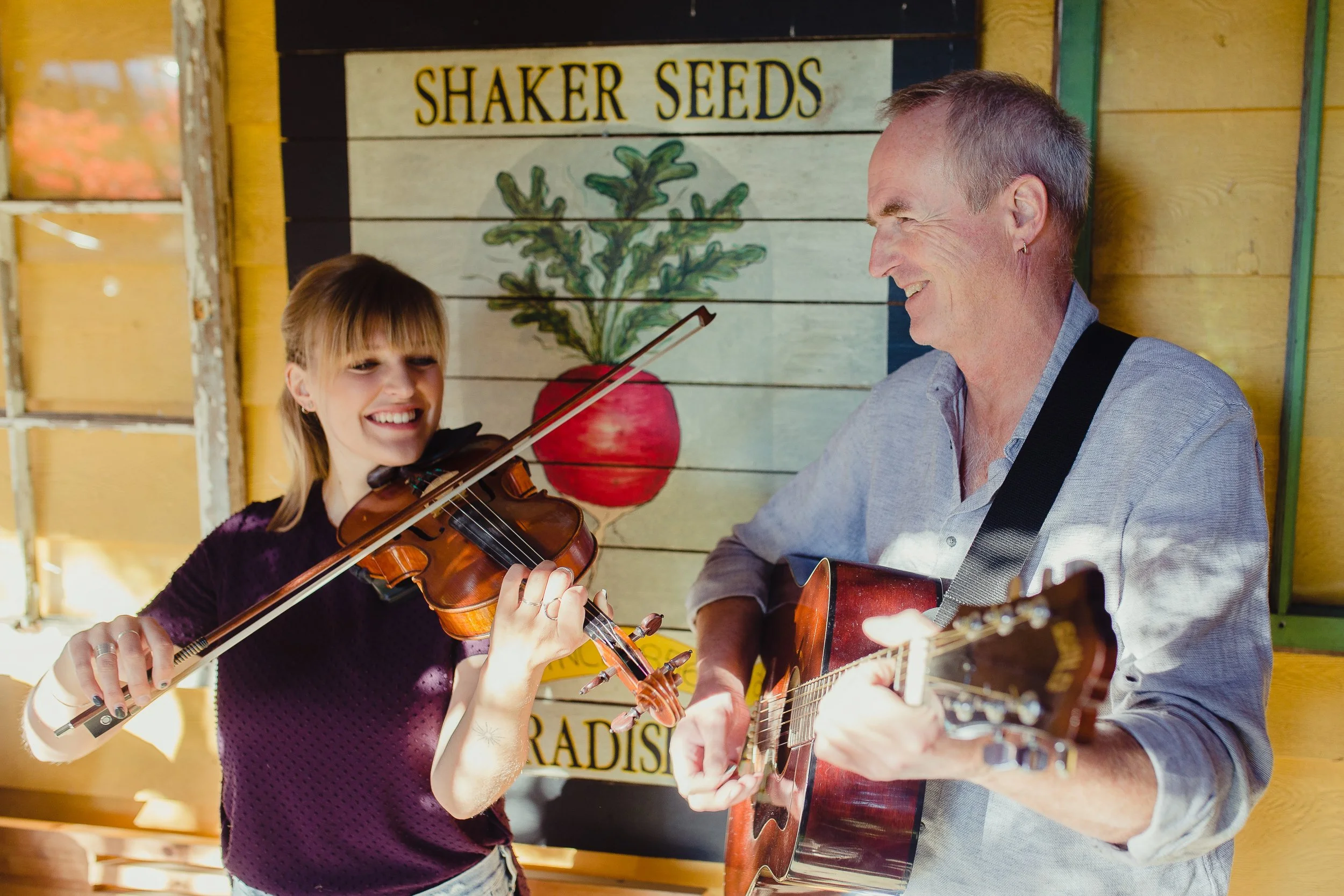 Ceilidh Briscoe, Victoria fiddle player, playing violin and Terry Boyle, Canadian singer songwriter, playing the guitar smiling at each other in front of a sign that says "Shaker Seeds" with a painted radish root and leaves.