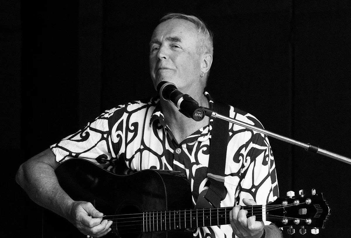 A man playing an acoustic guitar and singing into a microphone on stage in black and white.
